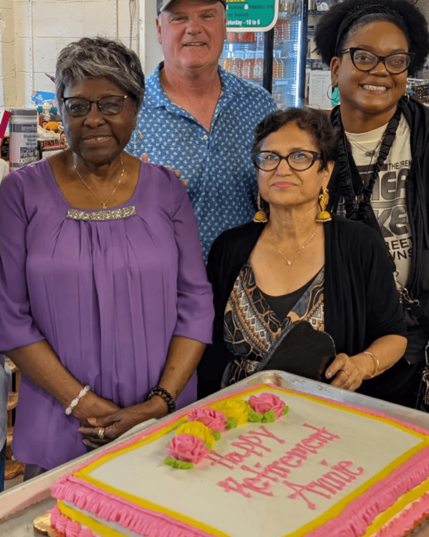 Four people standing behind a birthday cake with pink and yellow decorations, celebrating an anniversary.
