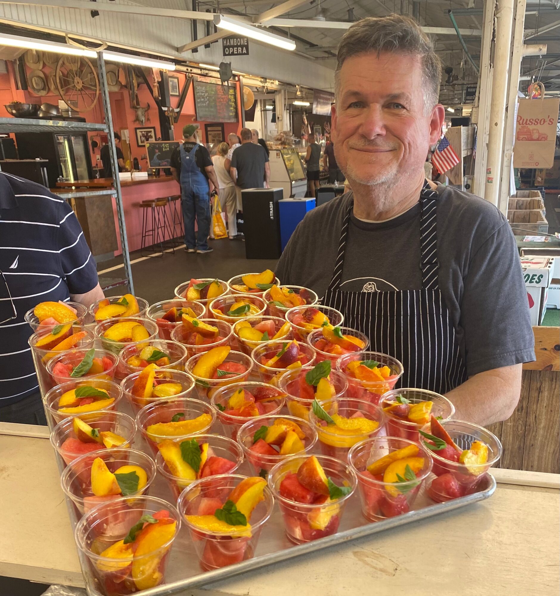 Smiling man in apron behind tray of fruit cups at market stall, with people and market setup in background.
