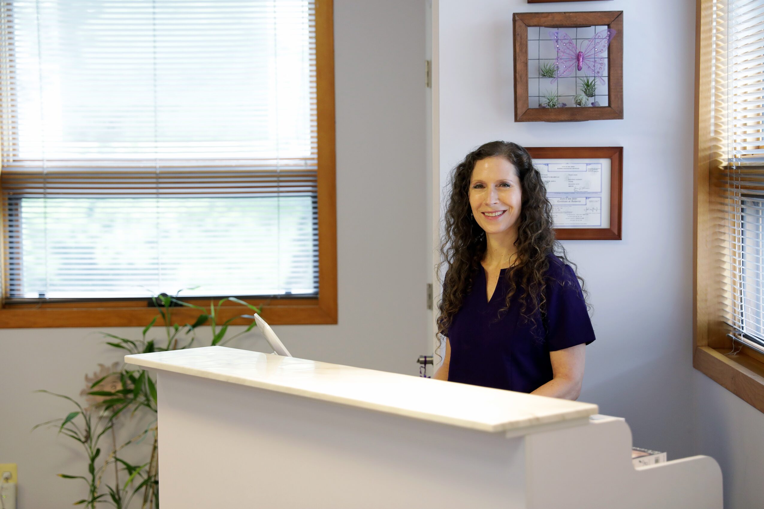 Woman with curly hair smiling in a room with windows and framed pictures on the wall.