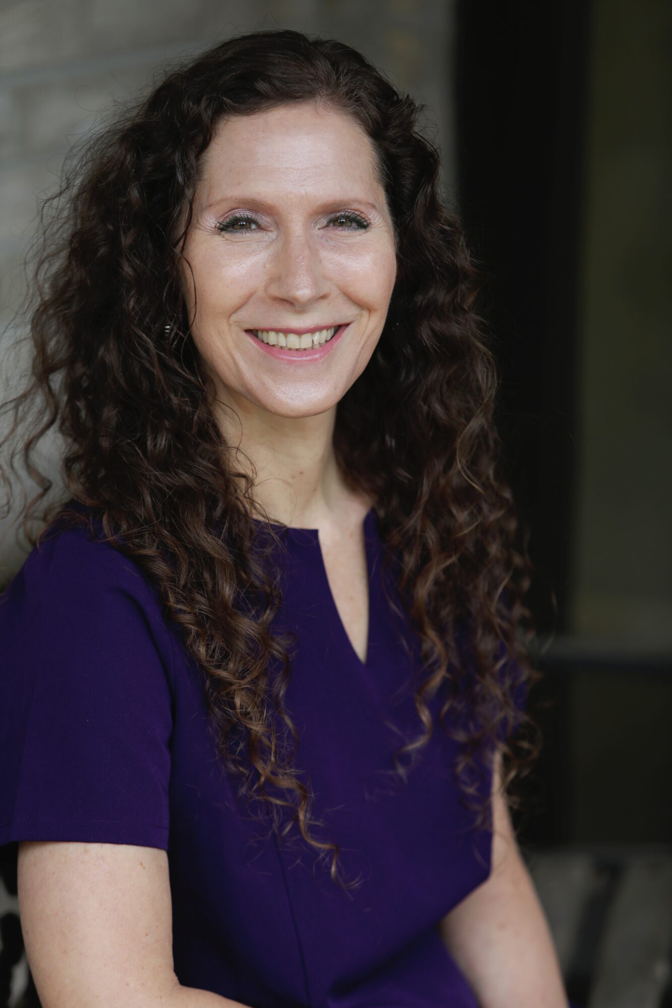 Smiling woman with long curly hair wearing a purple top, standing outdoors.