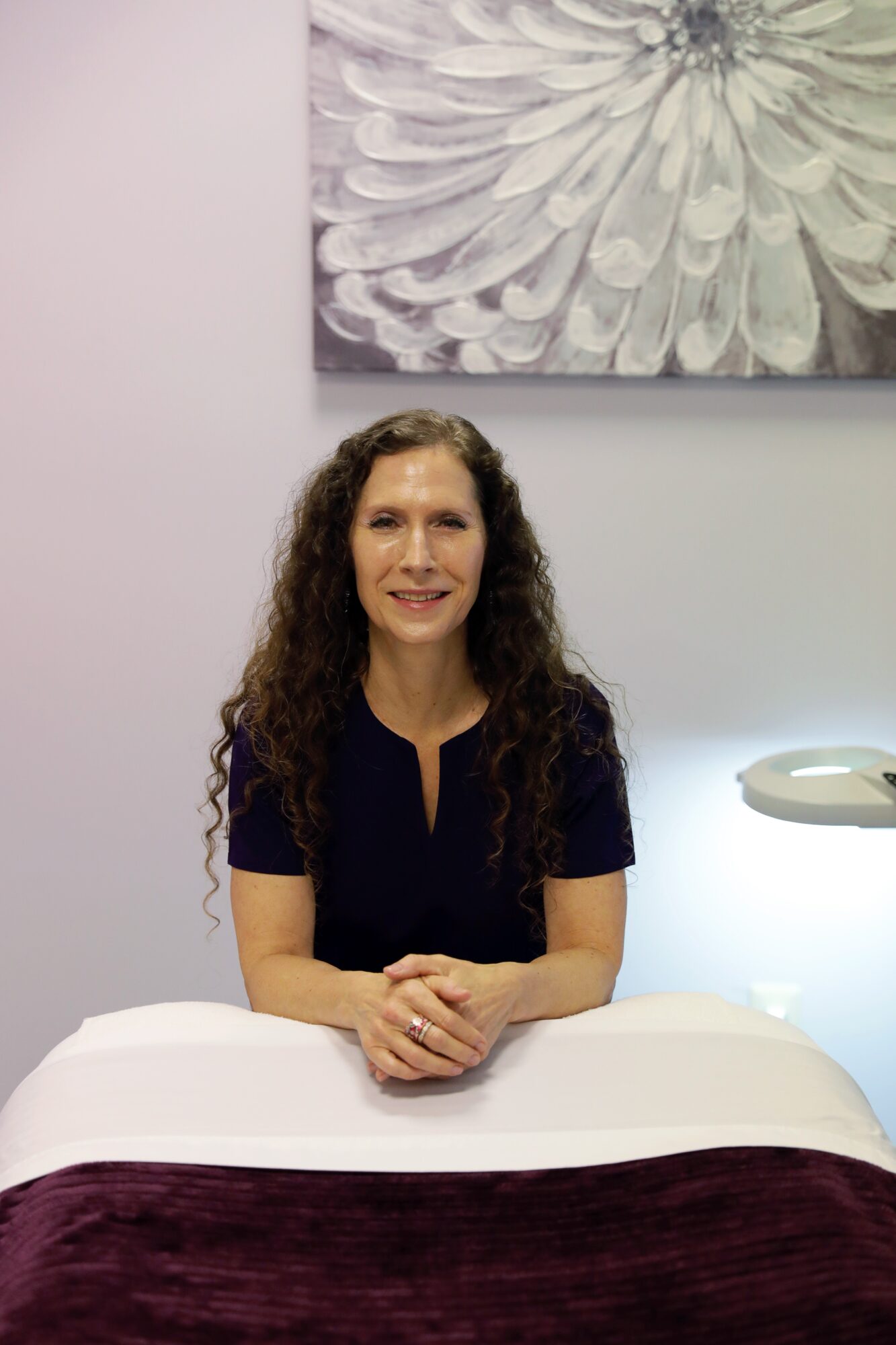 Woman with long curly hair sitting at a table, smiling, with a large flower painting above her and a lamp nearby.