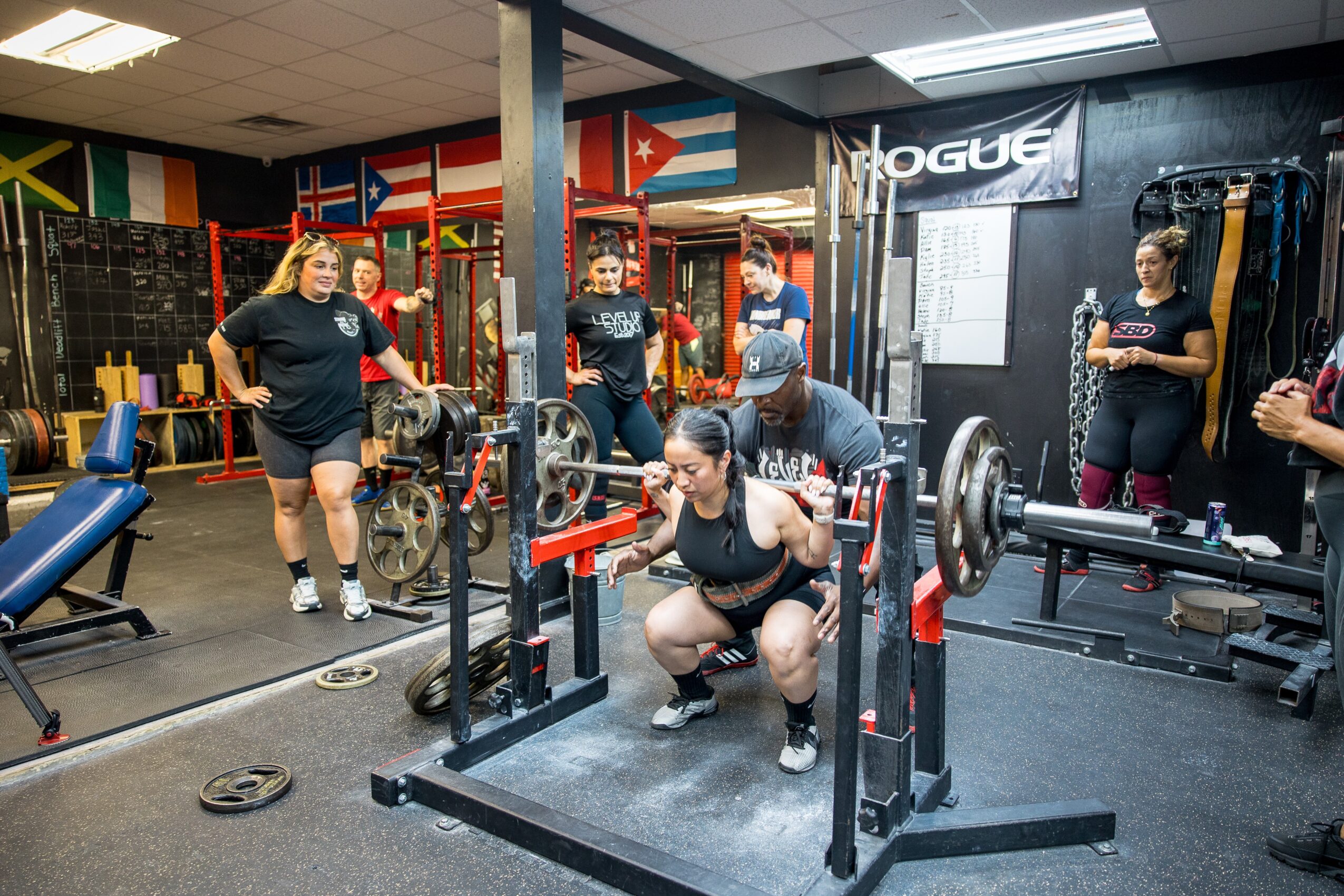 People in a gym with flags on the wall, some lifting weights, one person squatting with a barbell, others watching.