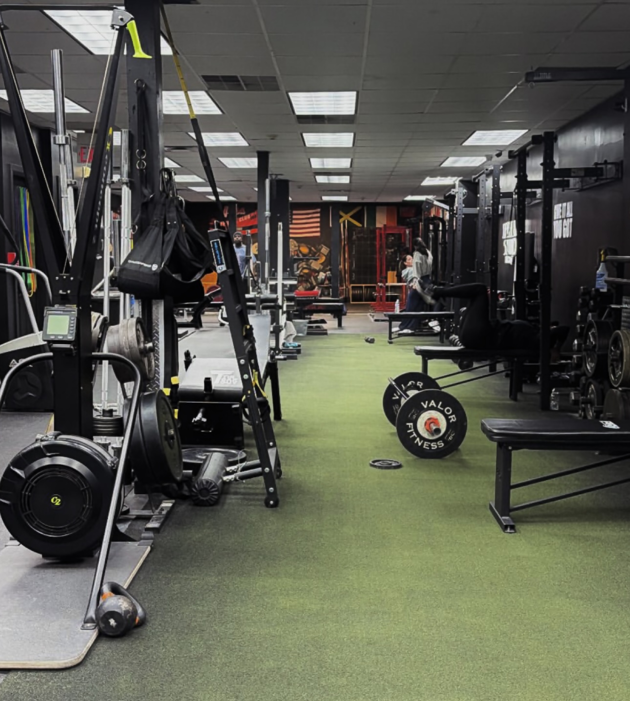 Empty gym with workout equipment, weights, and benches, illuminated by ceiling lights, with flags and gym equipment in the background.