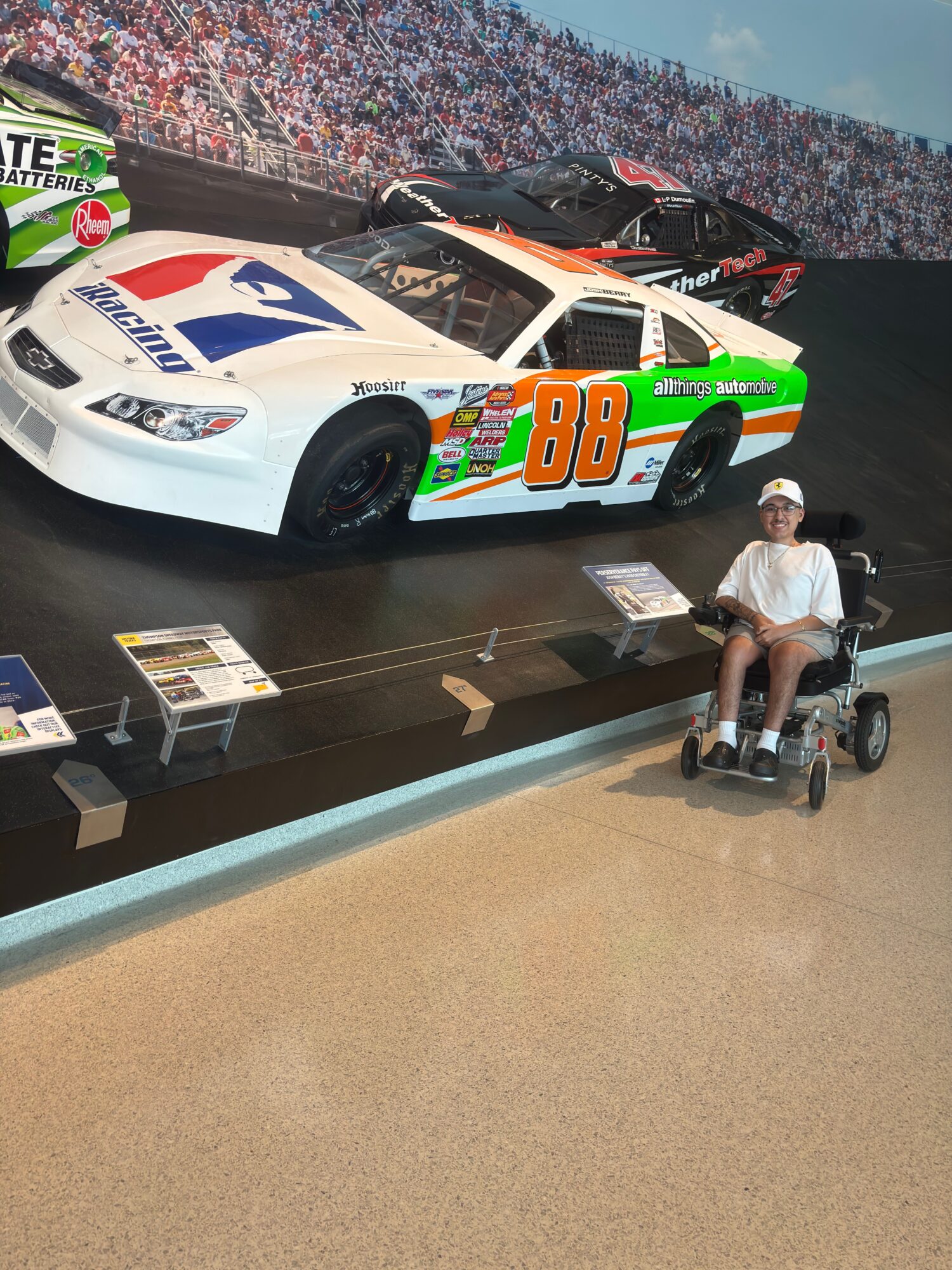 Person working on a race car in a garage, with a crowd in the background, and a person using a tool on the car.
