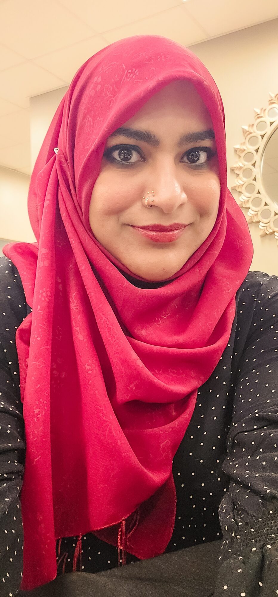 Woman wearing a pink hijab and black polka dot top, smiling indoors.