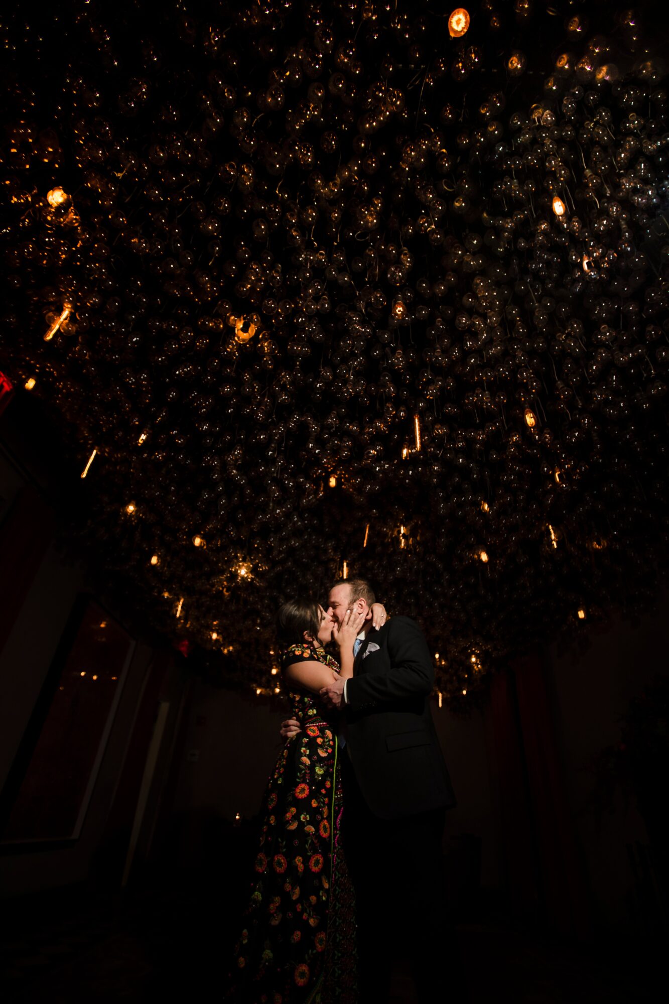 A couple dancing under a ceiling decorated with hanging lights, with a dark background and warm lighting.