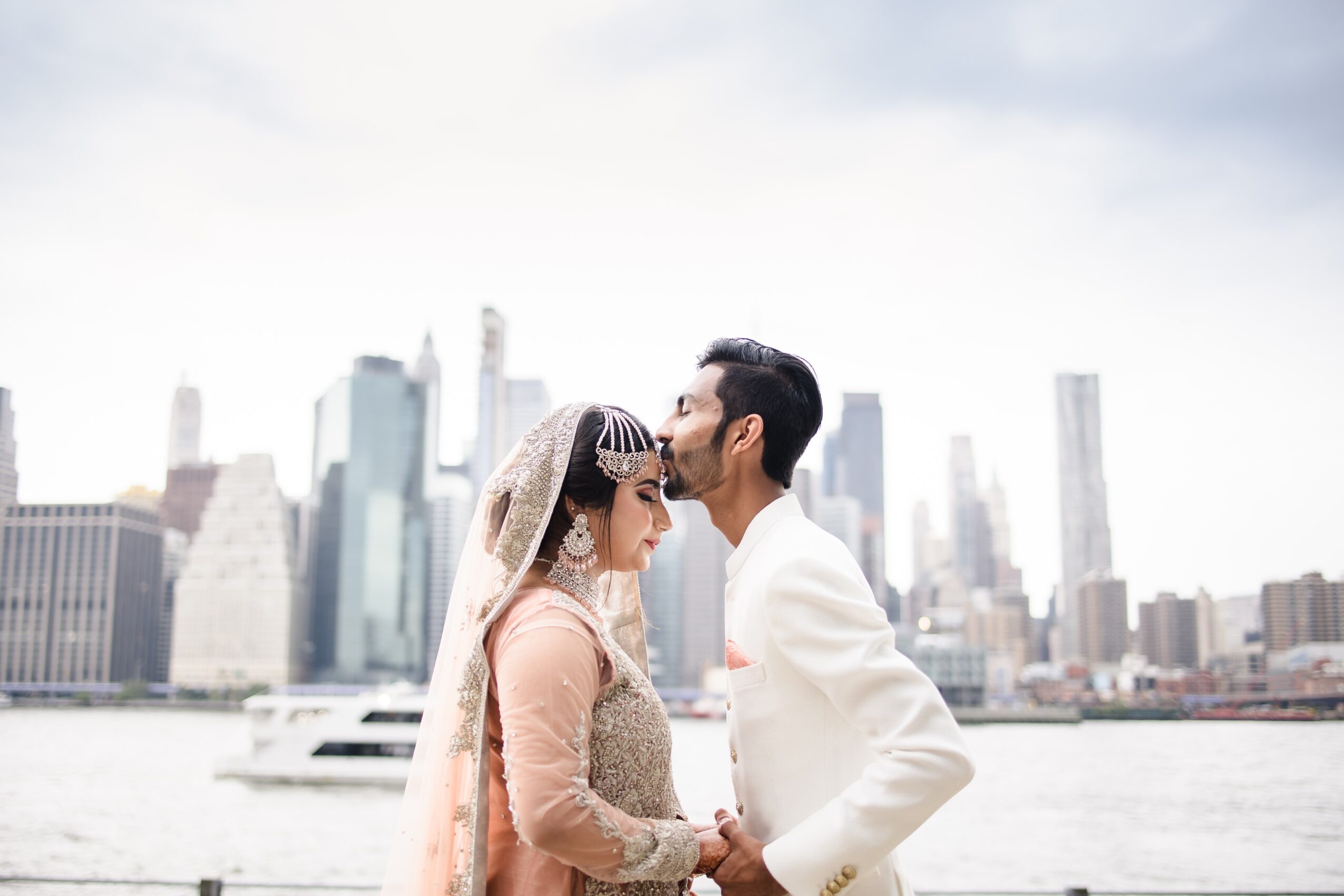 A couple in wedding attire face each other on a boat with a city skyline in the background.