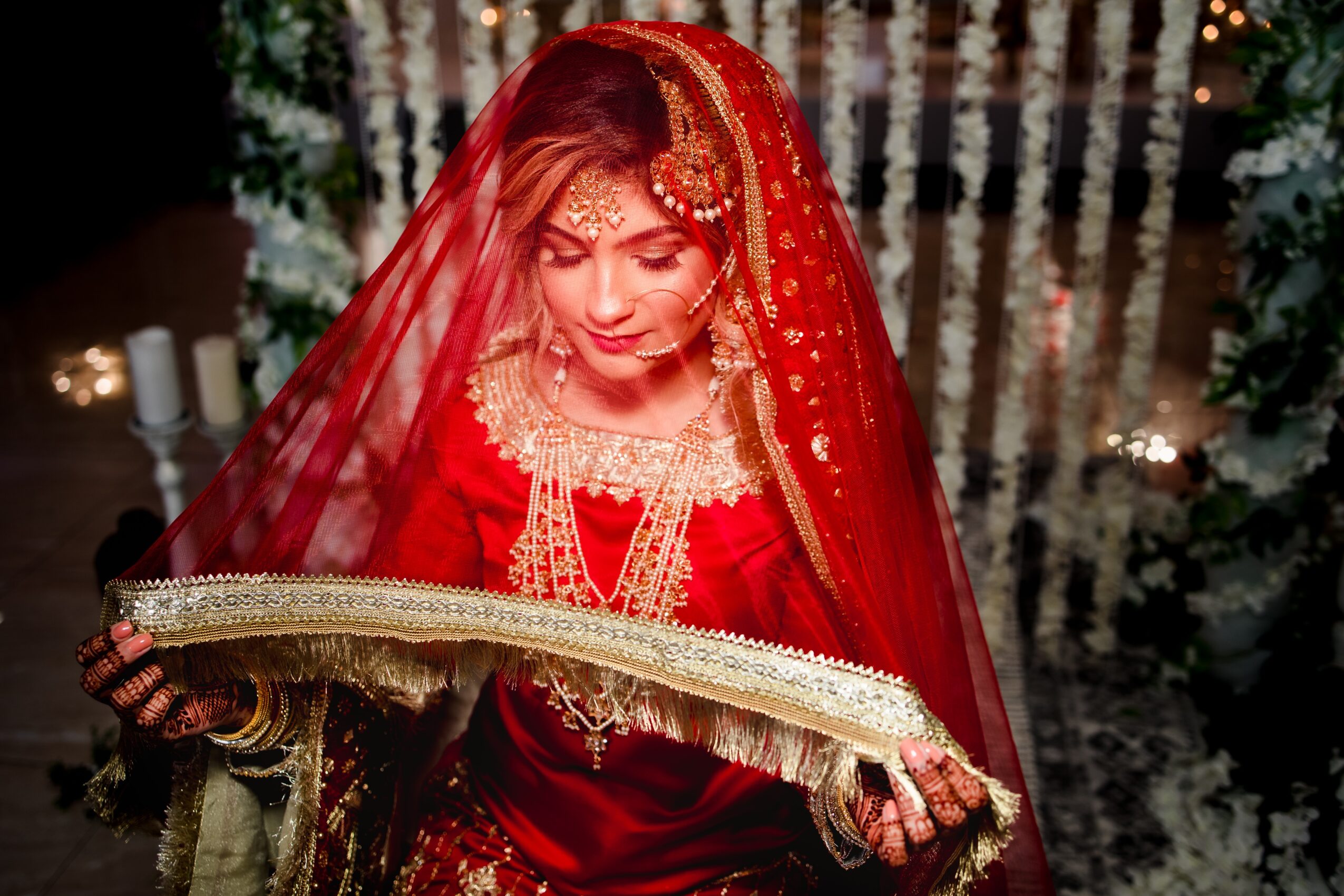 A woman in red traditional attire holding a decorative cloth, with a festive background of lights and greenery.