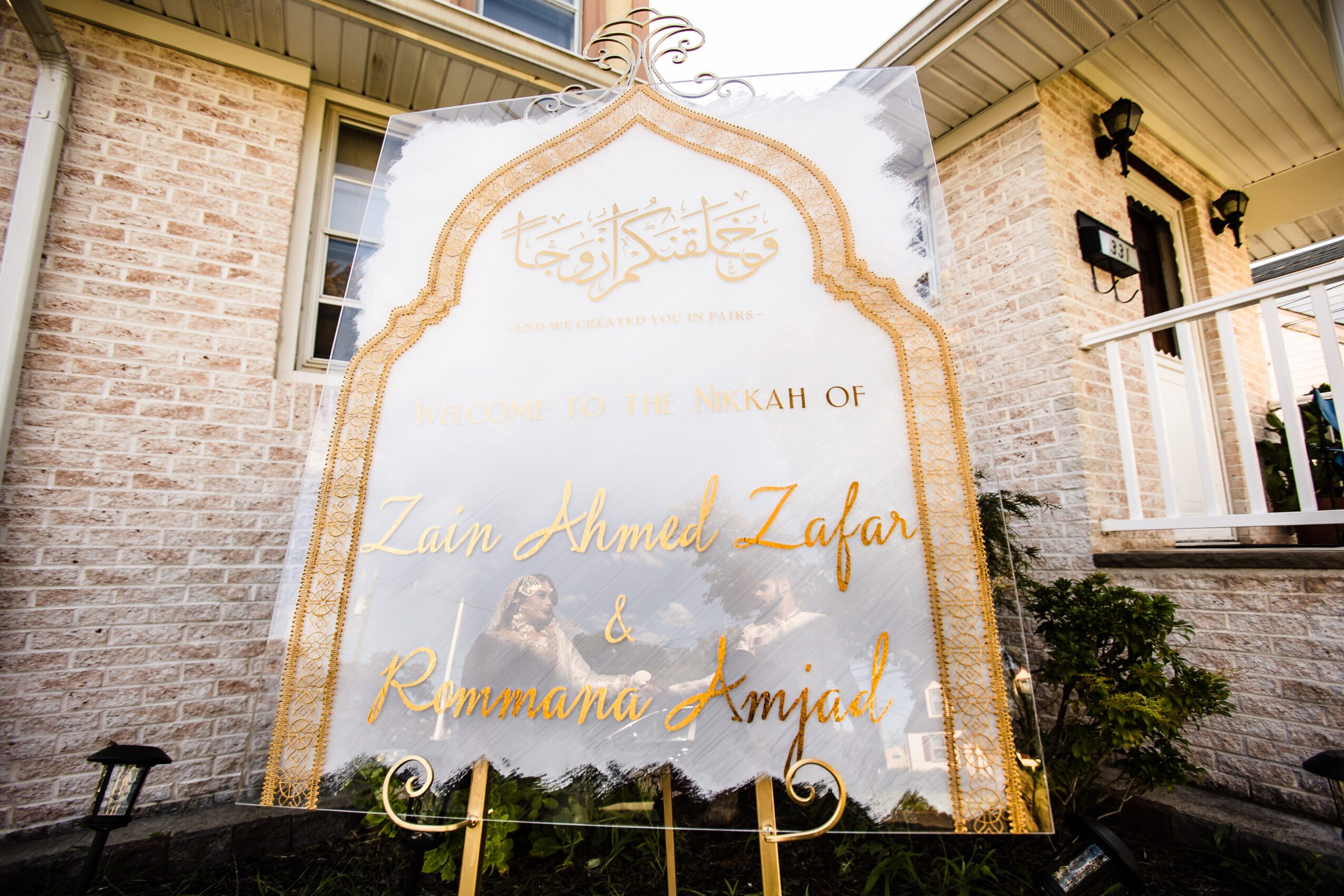 Decorative sign with Arabic script and names Zaan Ahmed Zafar and Rummana Amjad, outdoors with buildings and greenery in background