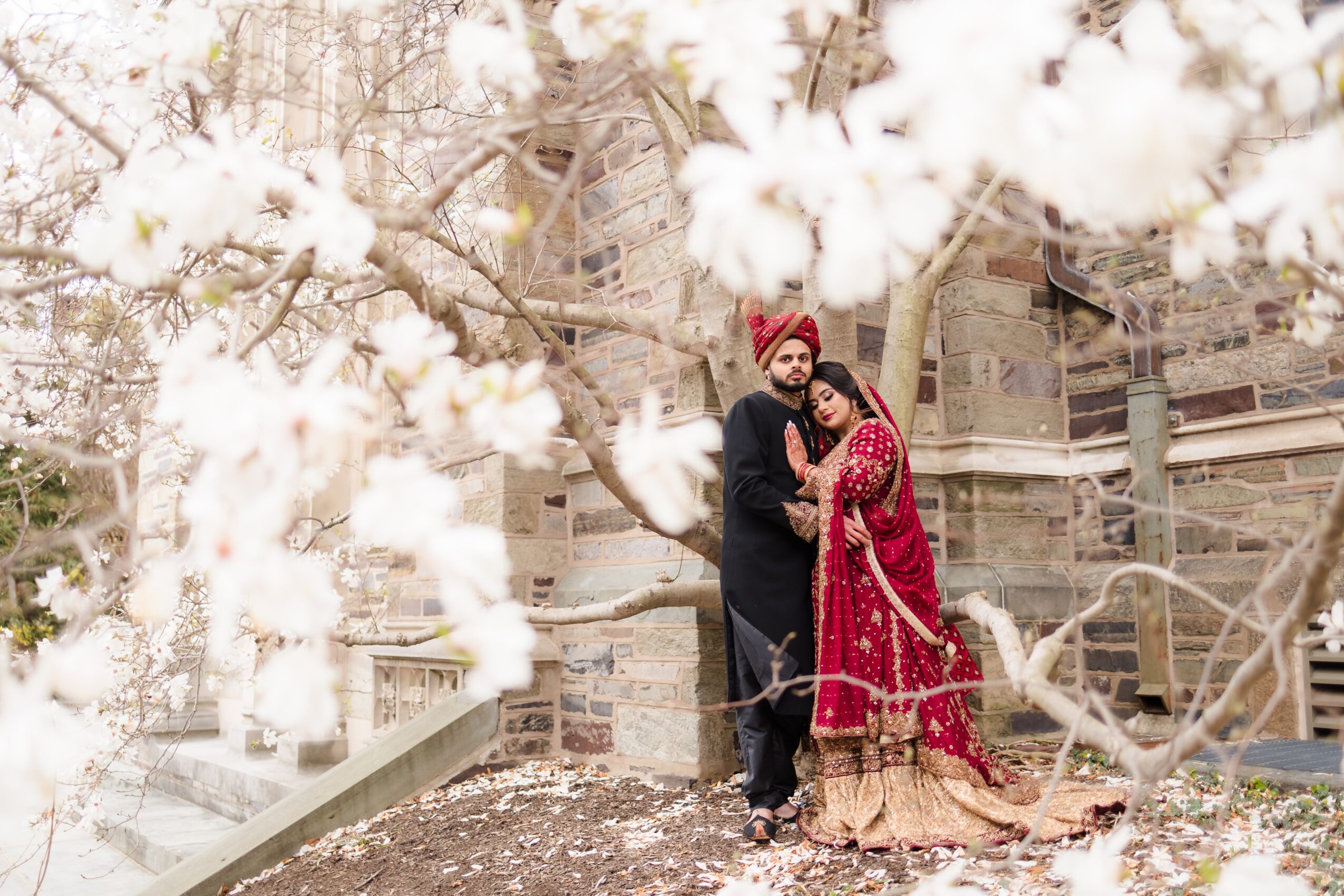 A couple dressed in traditional attire standing outdoors near a large tree with white blossoms, with buildings in the background.