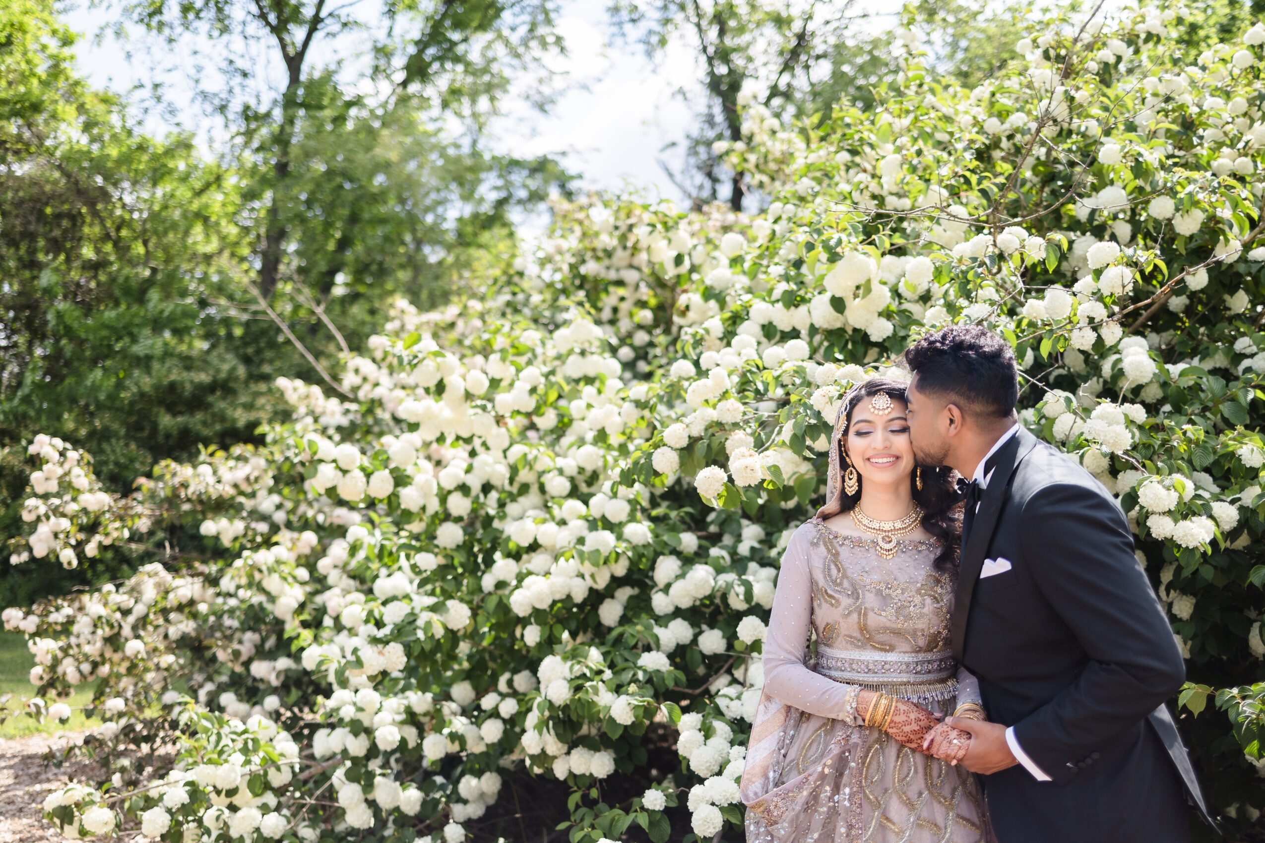 A couple stands in front of blooming white flowers, holding hands and smiling, outdoors on a sunny day.