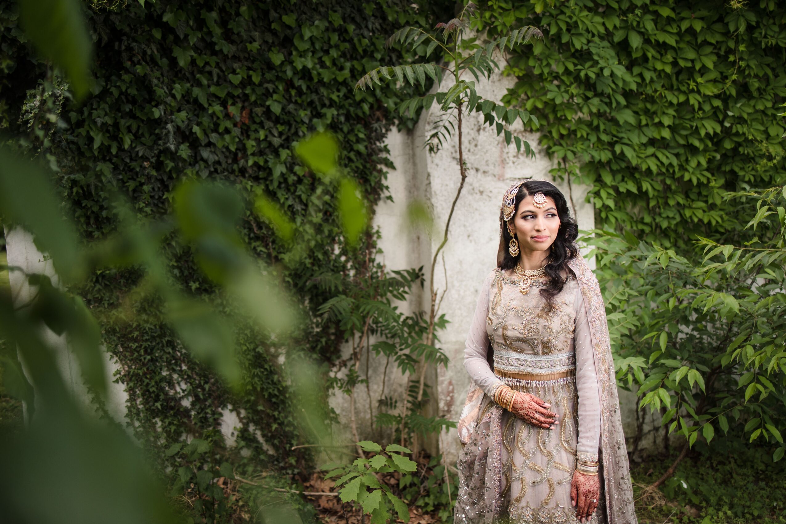 Woman in traditional attire standing outdoors near greenery and a white wall, looking to the side.