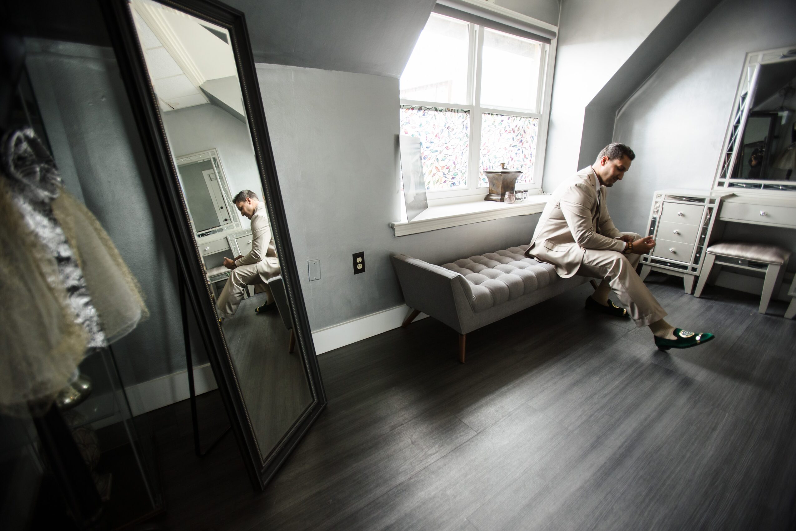 Person sitting on a bench in a room with a large window, a TV, and a mirror, with a reflection of the person.