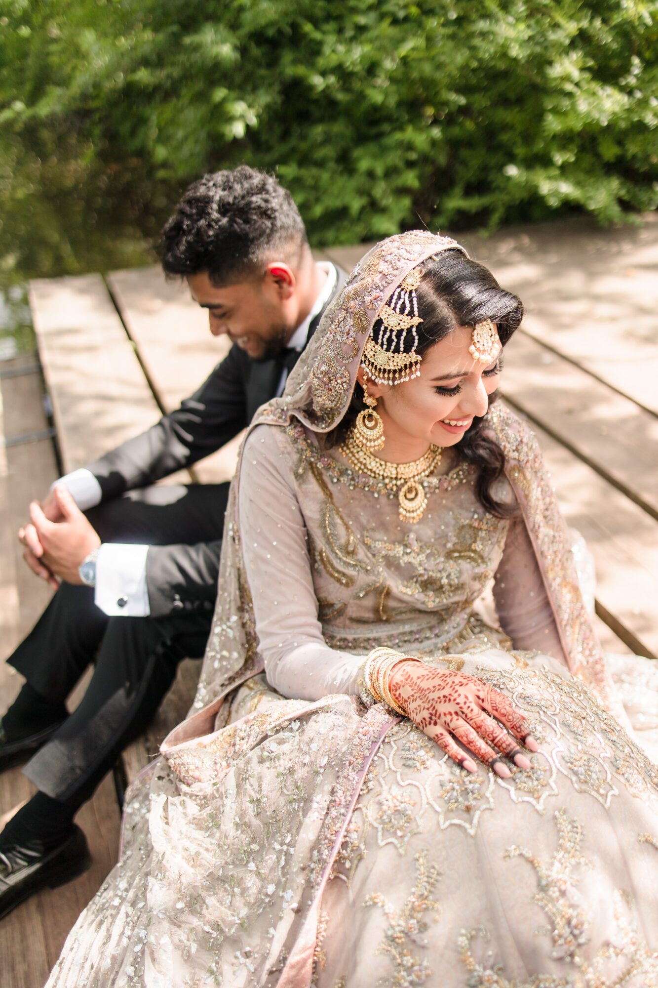 A bride and groom sit outdoors on a wooden bench, dressed in traditional wedding attire, with greenery in the background.