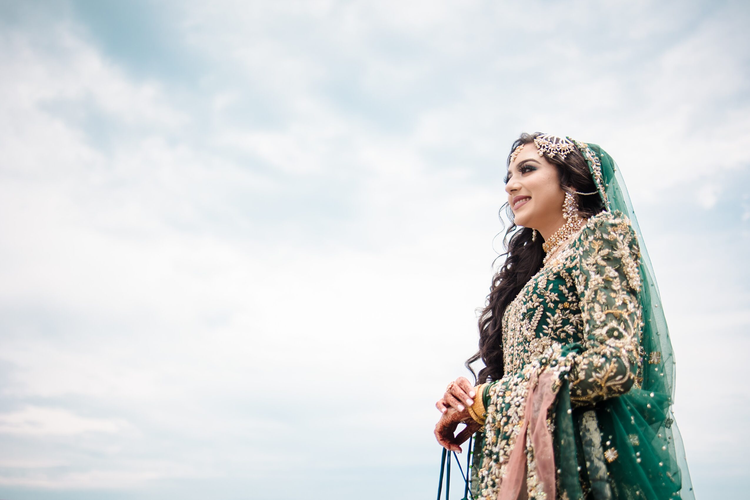 A woman in traditional attire stands outdoors against a cloudy sky, smiling and looking to the right.