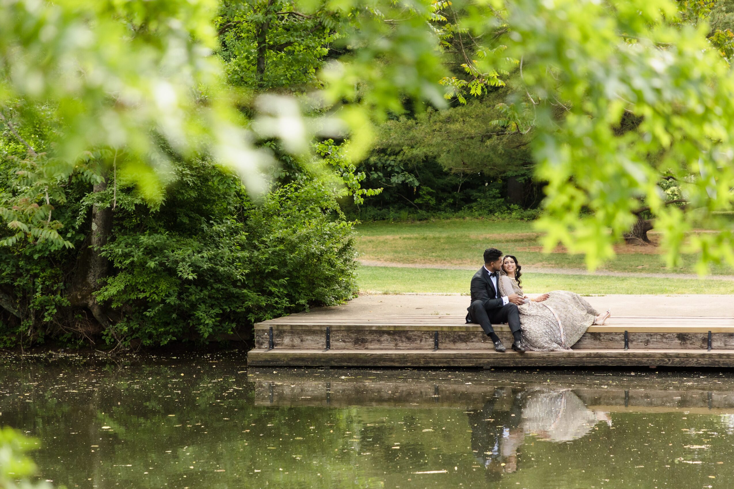 A couple sitting on a dock by a lake, surrounded by green trees, with their reflection visible in the water.