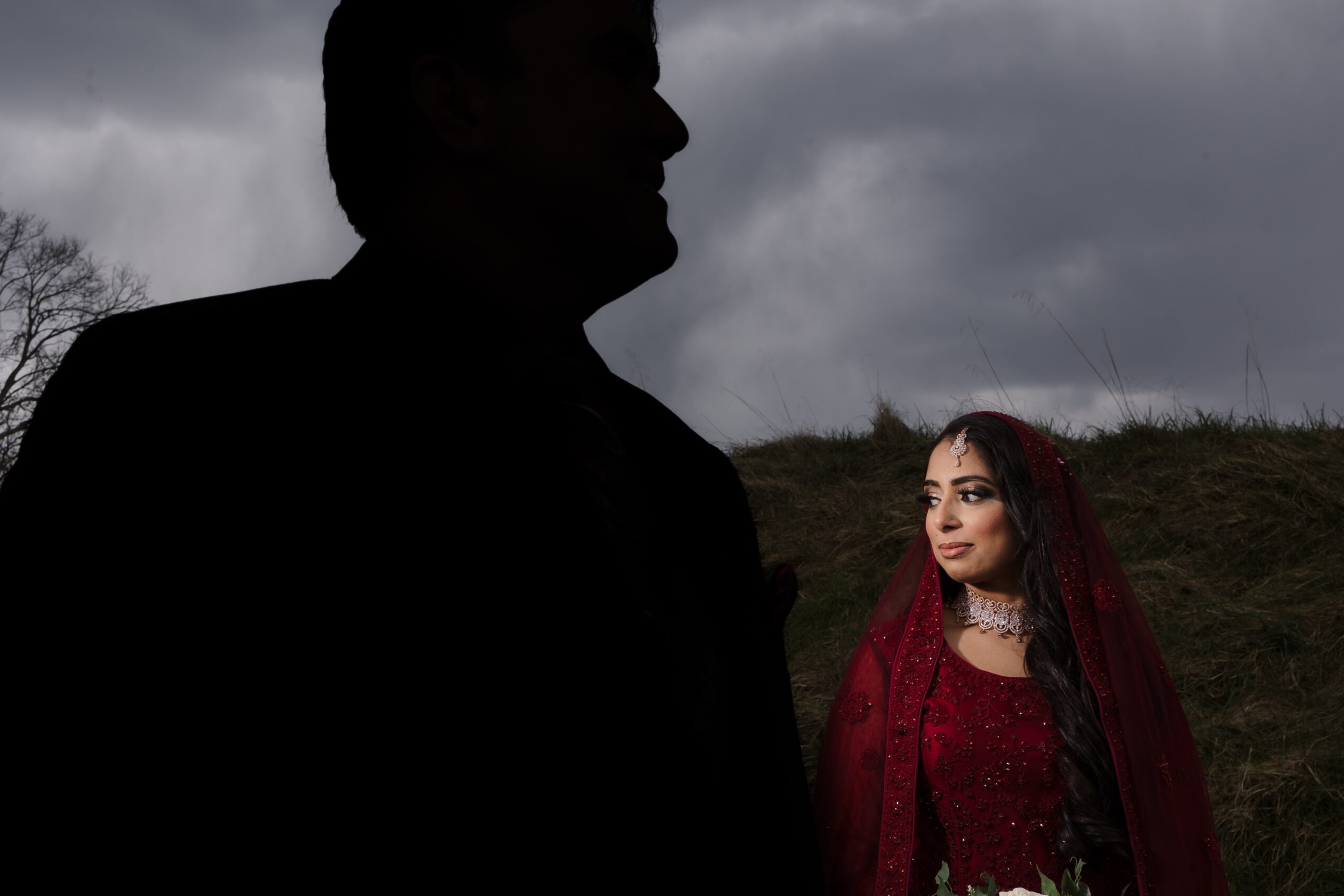 A woman in red traditional attire with jewelry stands outdoors, with a dark silhouette of a person in the foreground against cloudy sky.