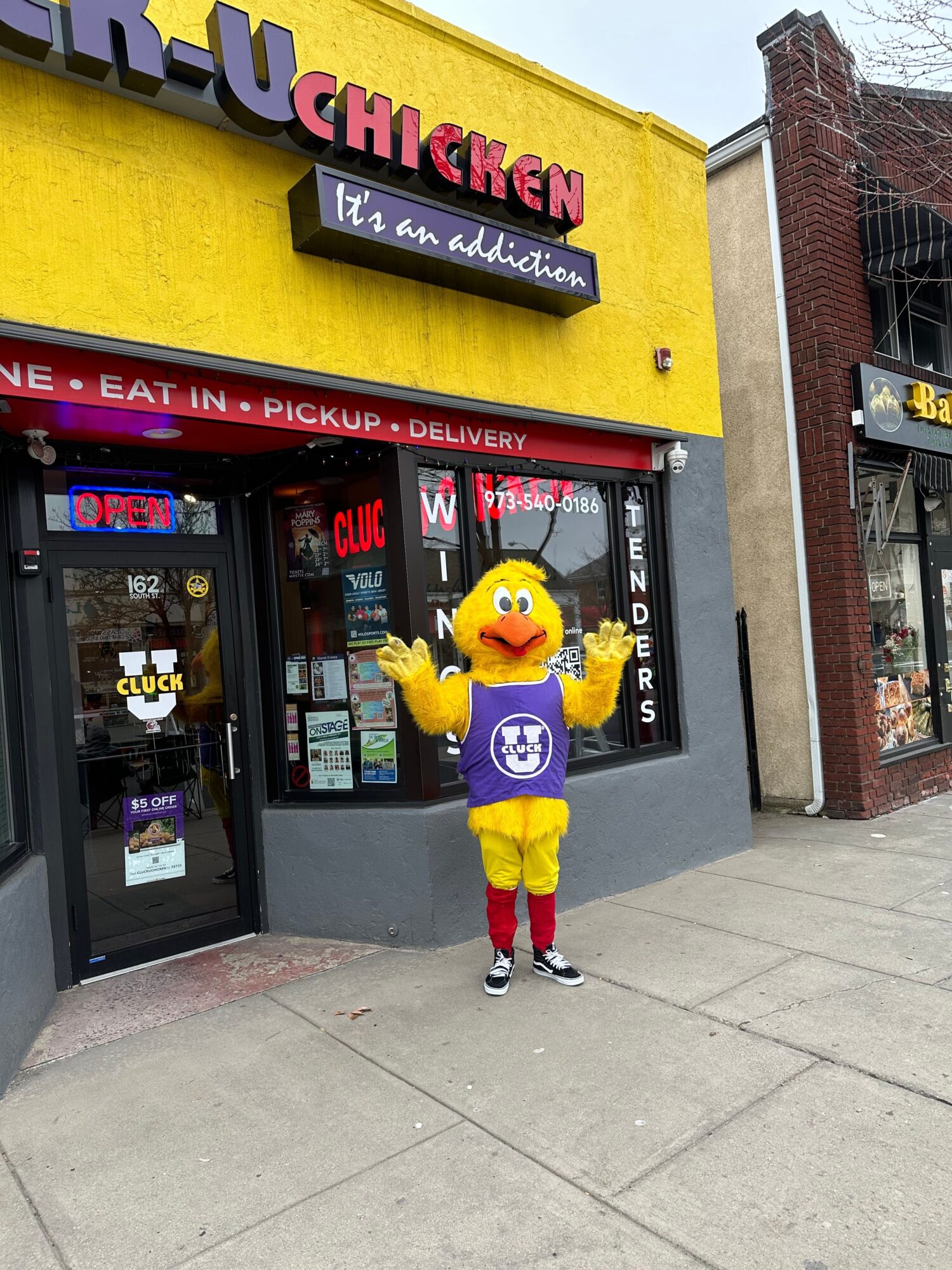 Person in a yellow mascot costume with a purple logo, standing outside a restaurant with a yellow sign, on a sidewalk.