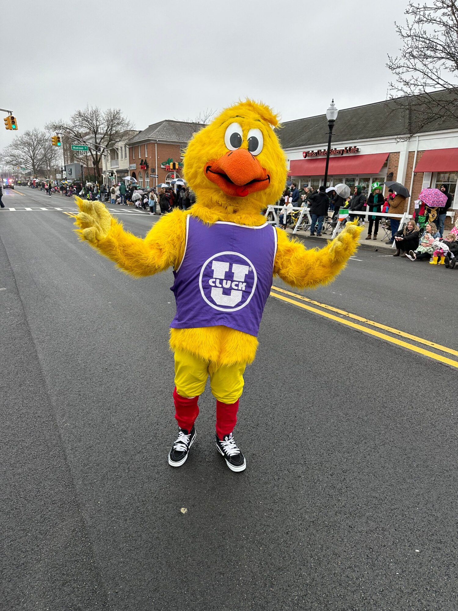 Person in a yellow bird mascot costume with a purple shirt, standing on a street with buildings and people in the background.