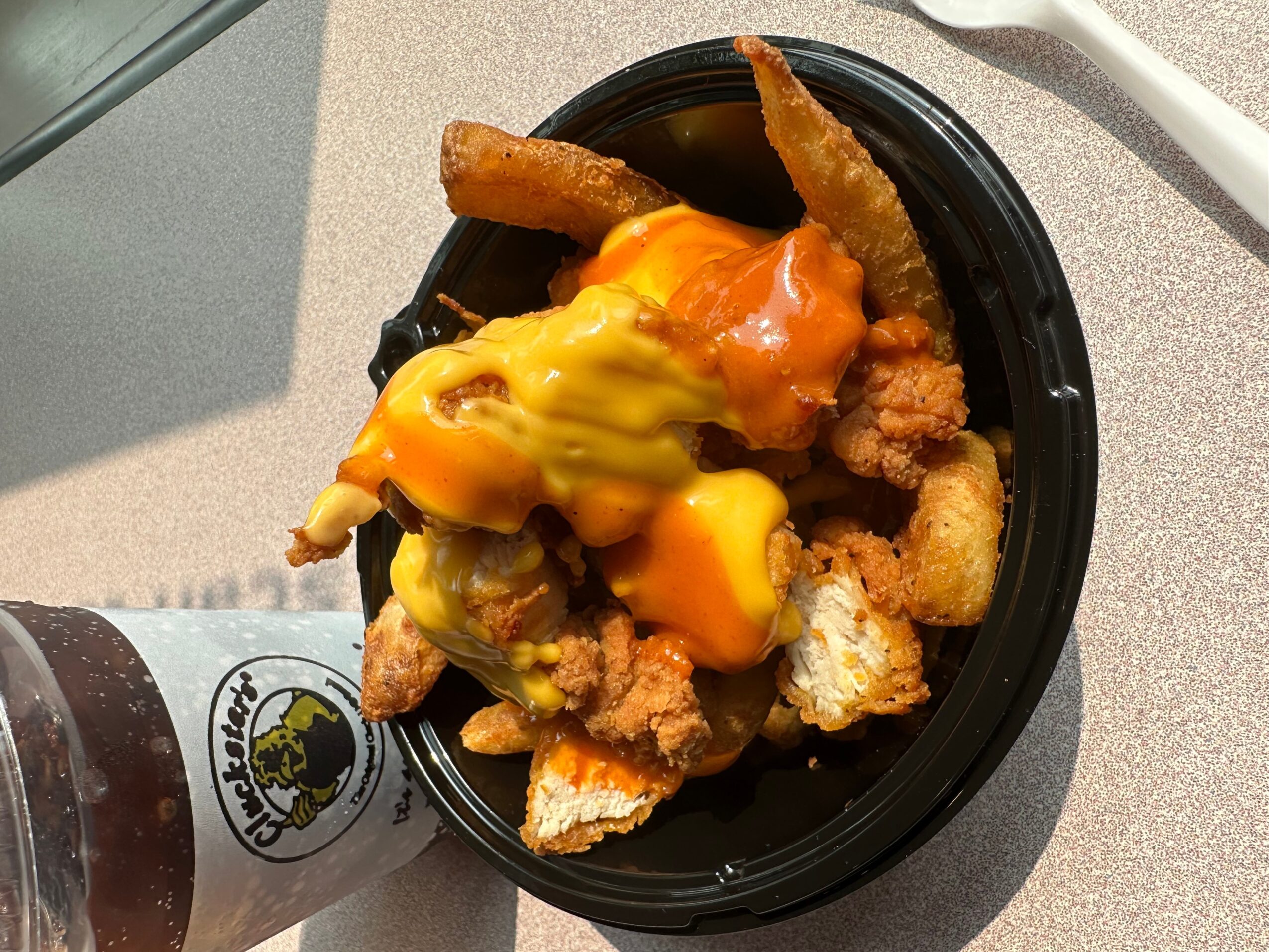 Fried chicken pieces topped with melted cheese and sauce in a black container, with a drink bottle nearby.