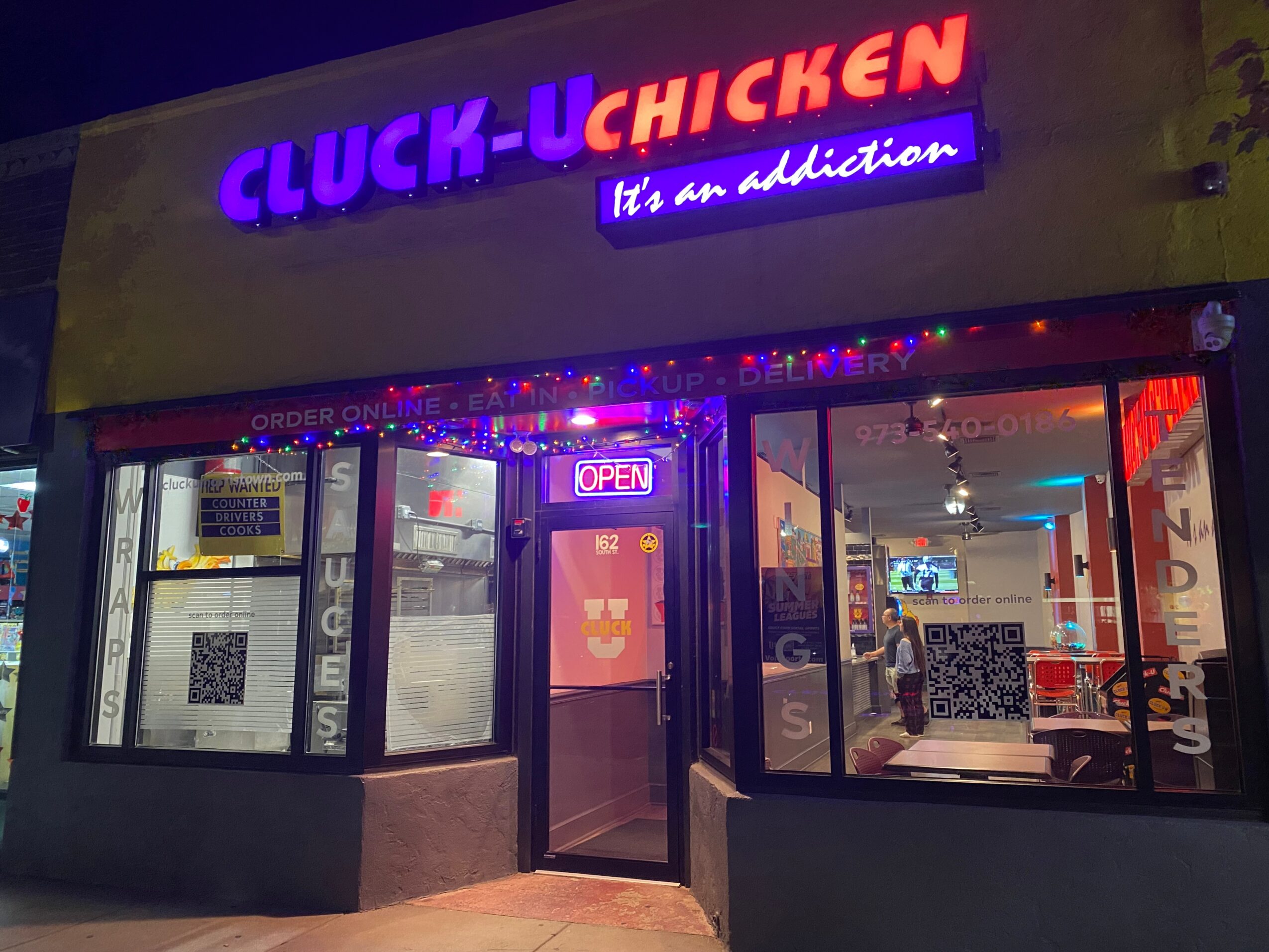 Night view of a restaurant with bright neon signs, glass windows, and an illuminated interior, with a person inside. Word count: 20