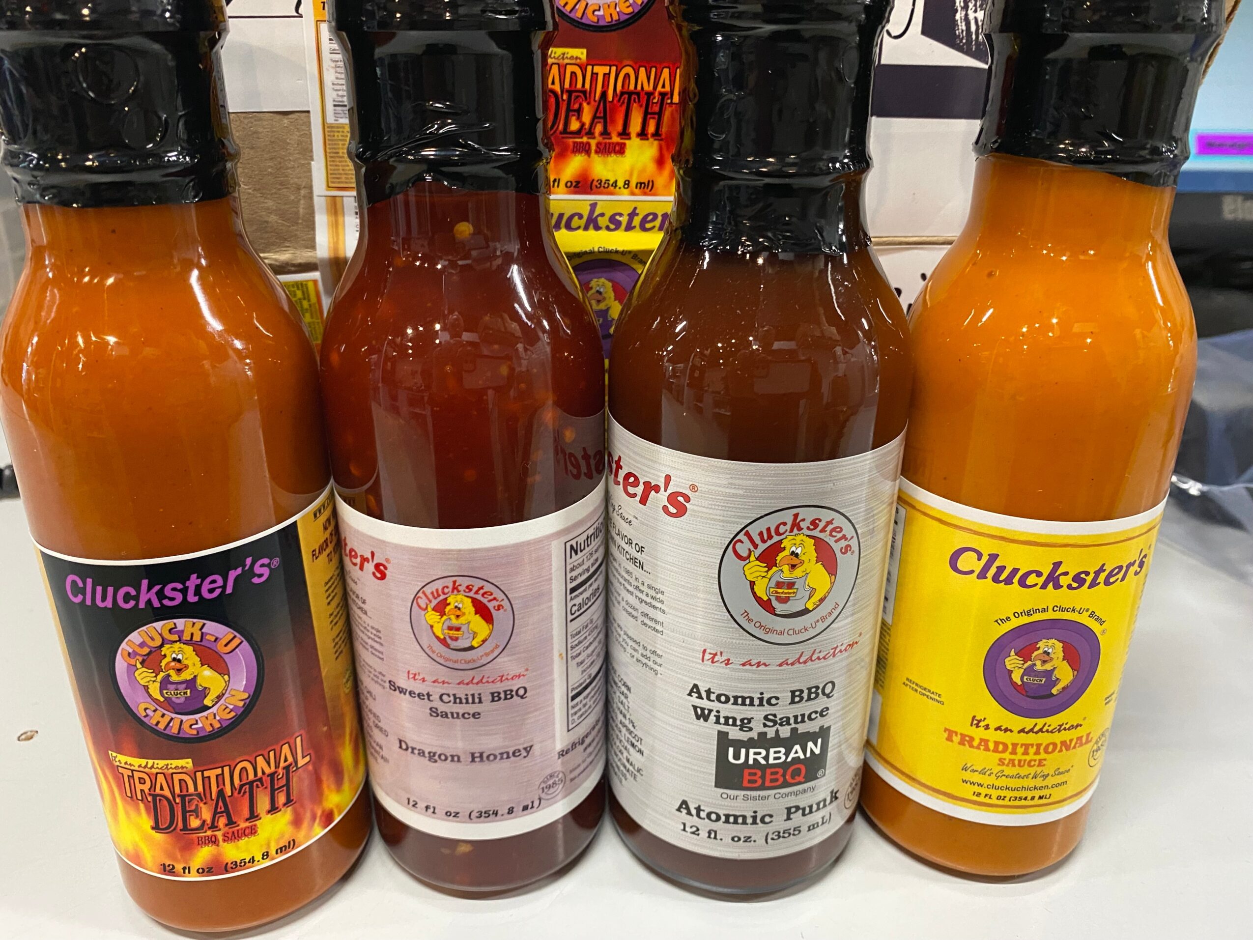 Four bottles of hot sauce with labels, two with black caps, on a shelf, with a yellow and purple label on the right.