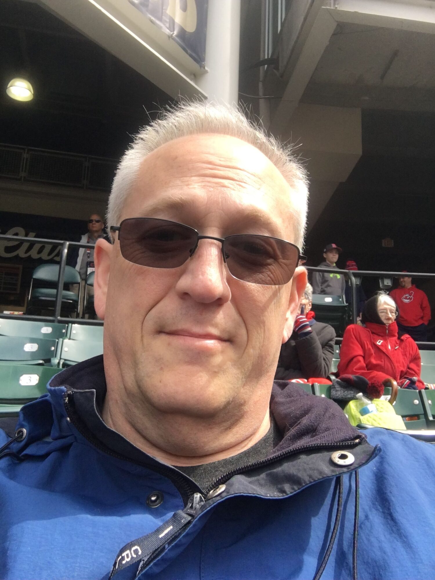 Man with gray hair wearing sunglasses and a blue jacket, smiling outdoors with people and red objects in background.