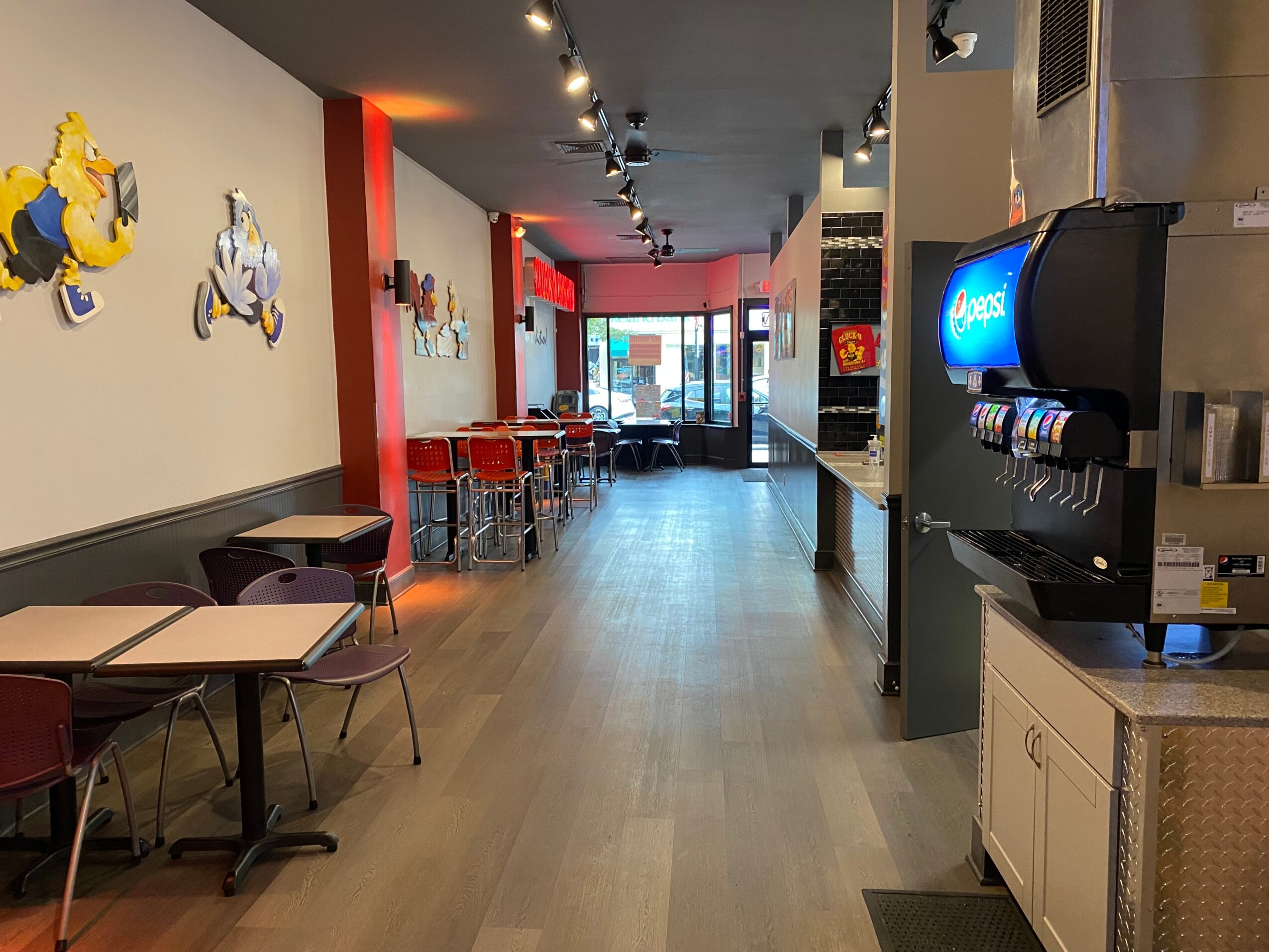 Empty restaurant with tables, chairs, wall decorations, and a beverage dispenser, illuminated by ceiling lights and natural light from the entrance.