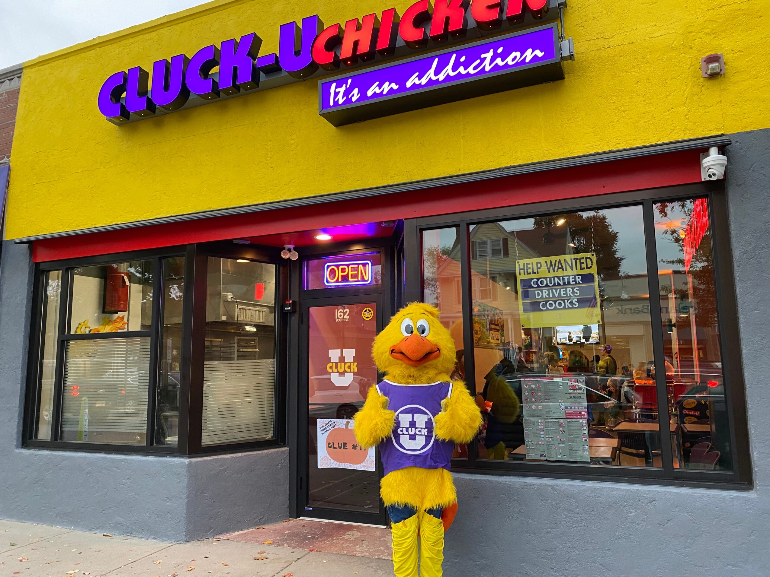 Yellow chicken mascot with purple accessories standing outside a fast-food restaurant with colorful signs and windows.