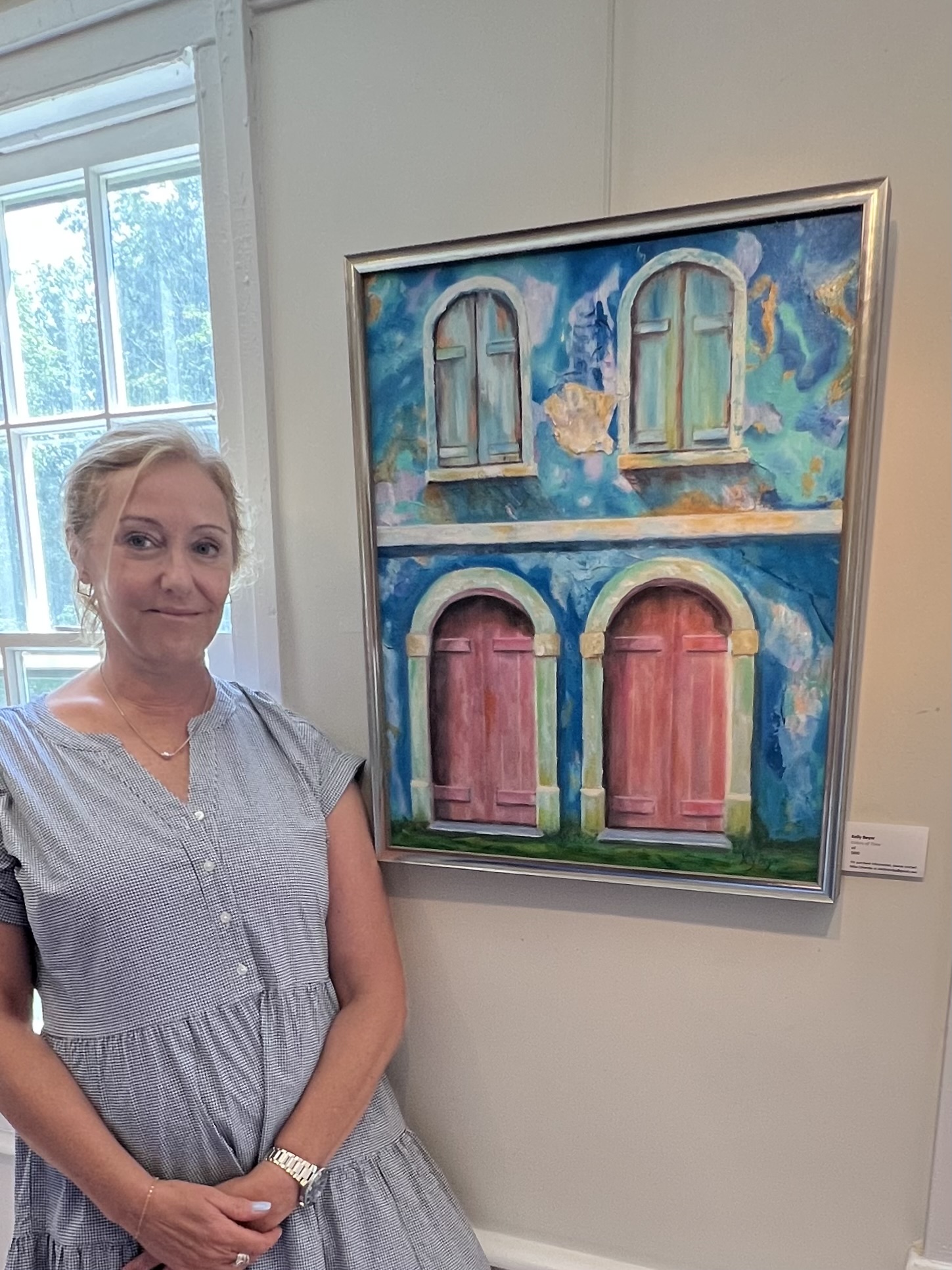 Woman with blonde hair in a light gray dress standing next to colorful painting of windows and doors on a white wall.