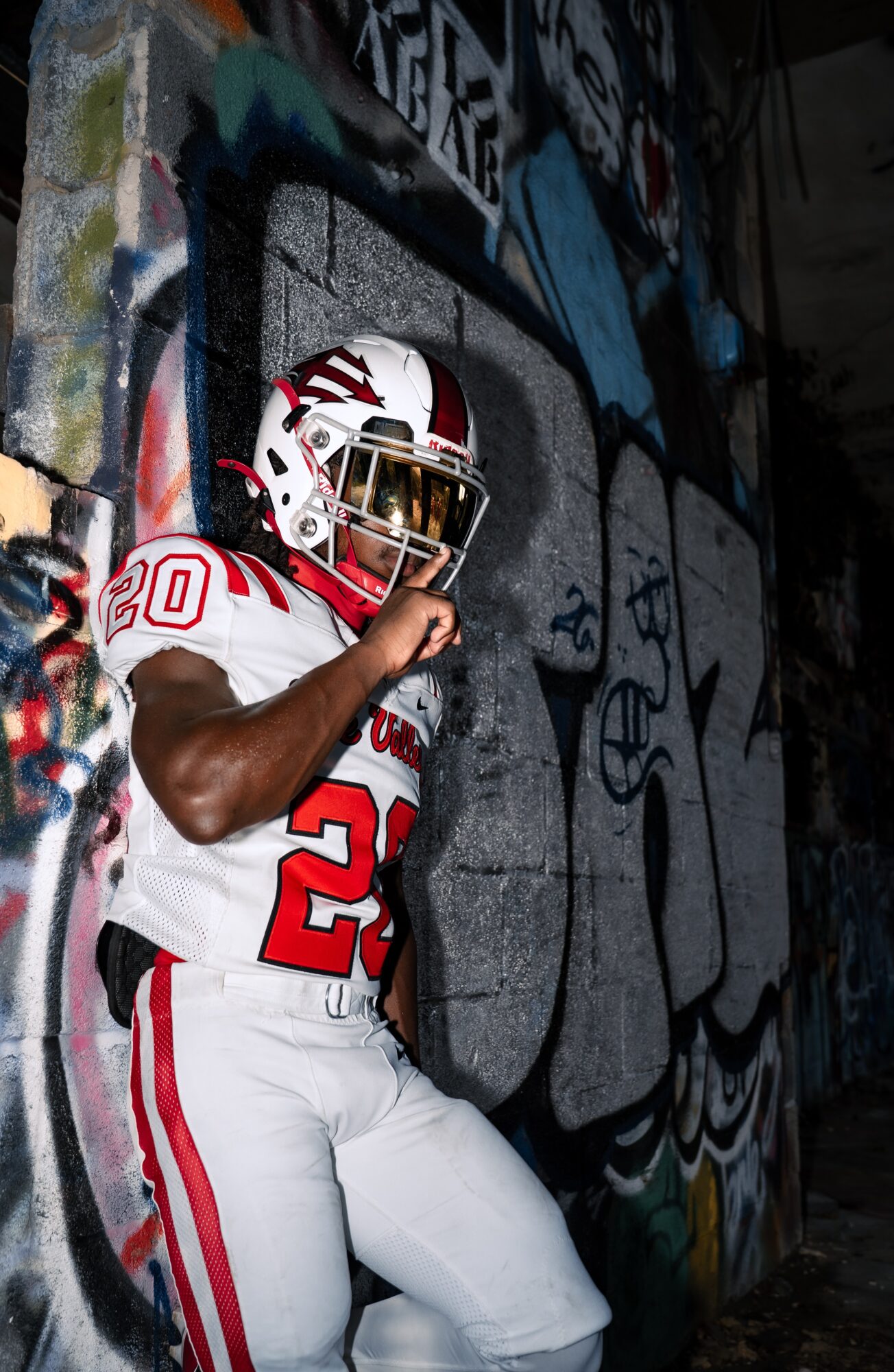 Football player in white and red uniform standing against graffiti-covered wall, holding helmet and looking to the side.