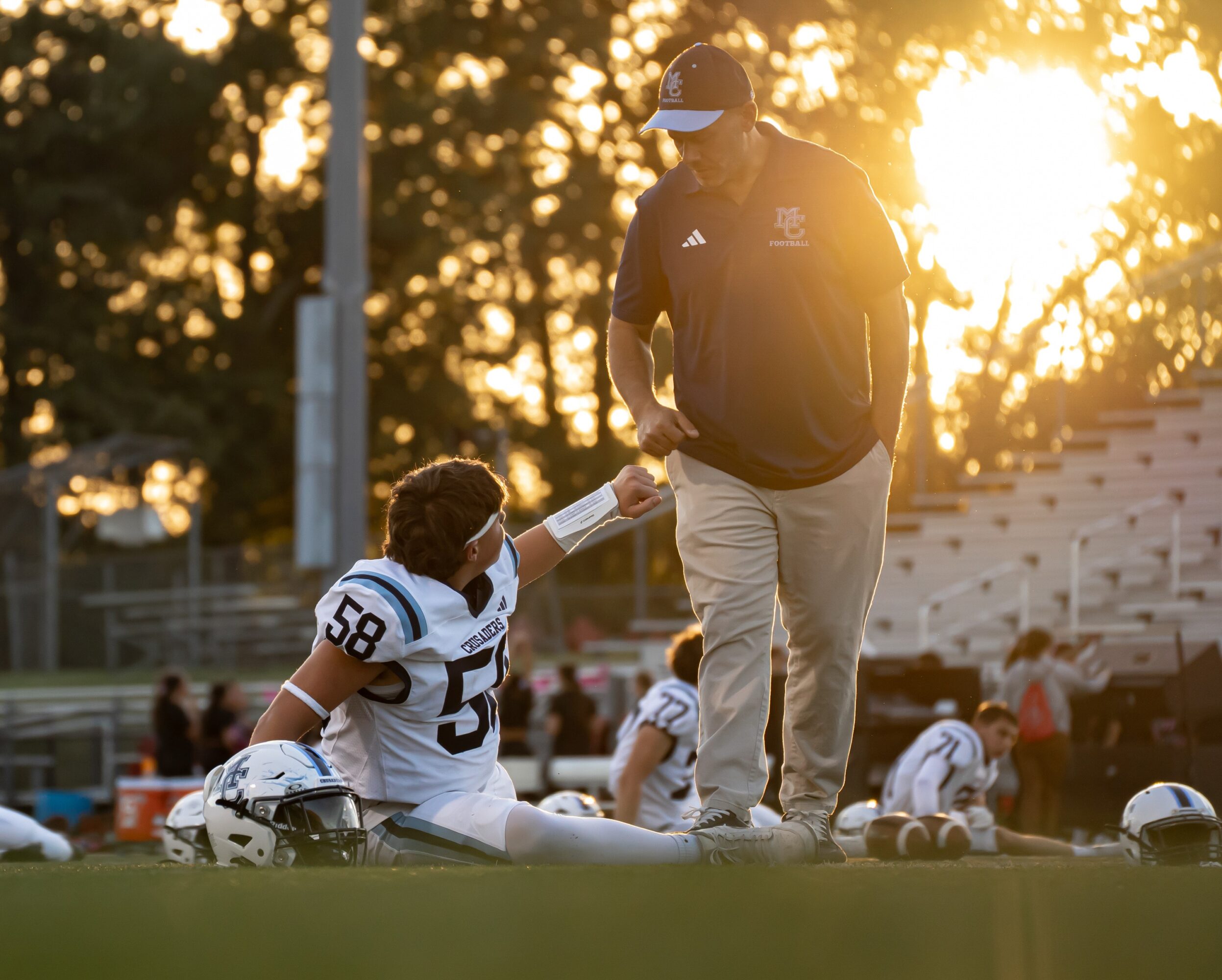 A football player sitting on the field receives water from a coach during sunset, with other players in the background.