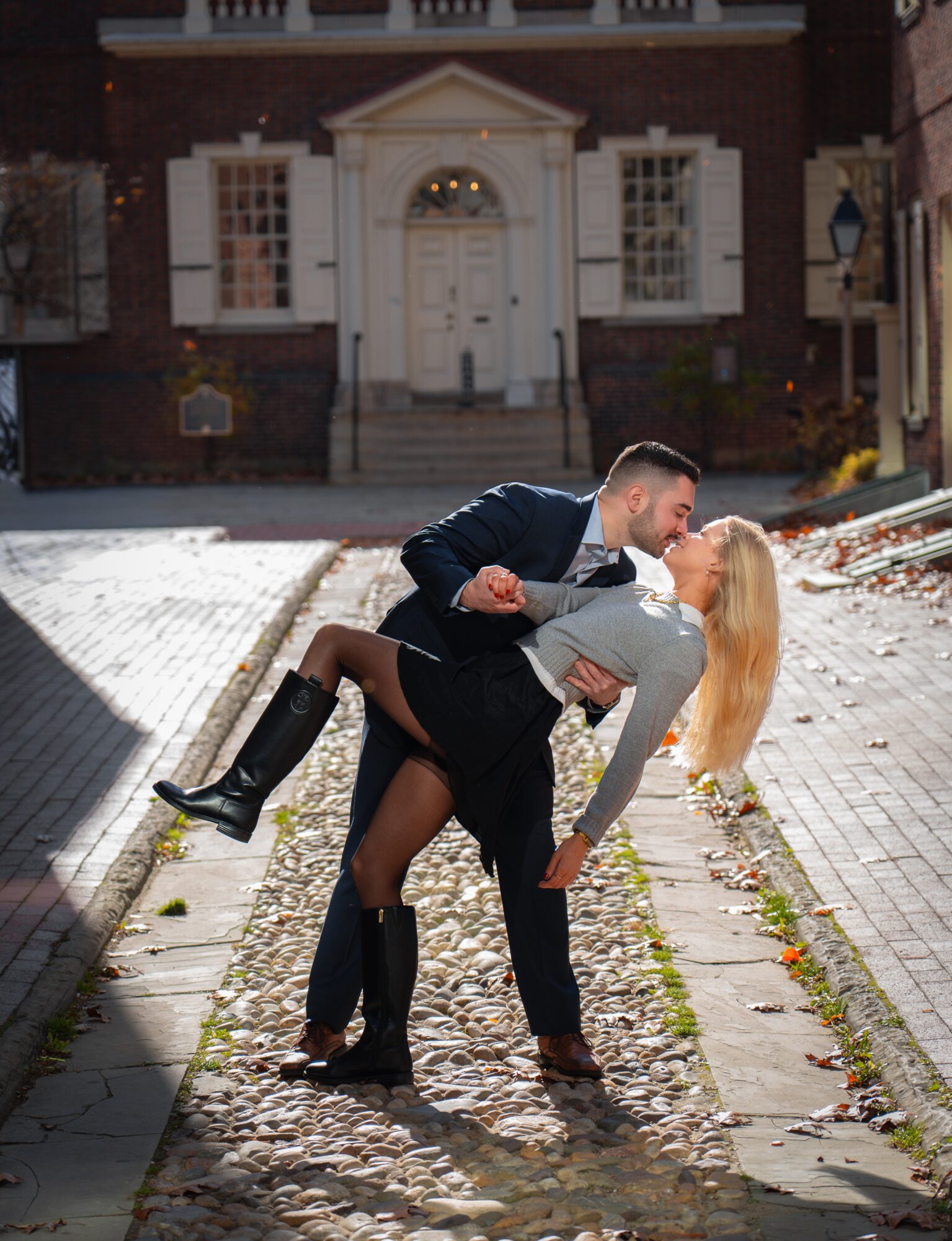 Couple kissing on cobblestone street in front of a large brick house with white door and windows, sunny day.