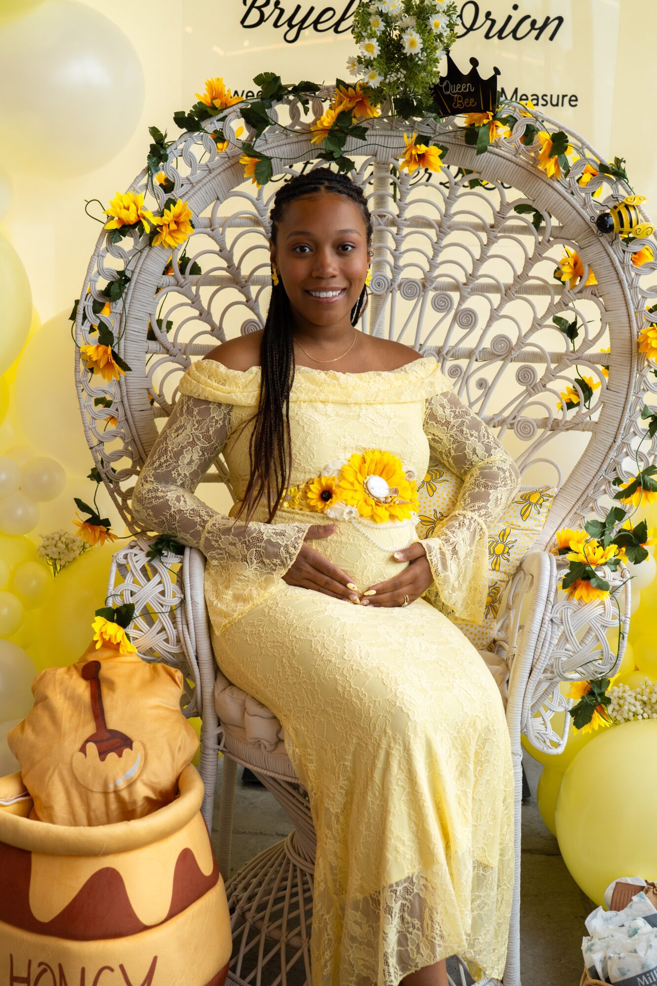 Young woman in yellow dress sitting on decorative white chair with floral and pumpkin decorations, smiling, holding a bouquet.