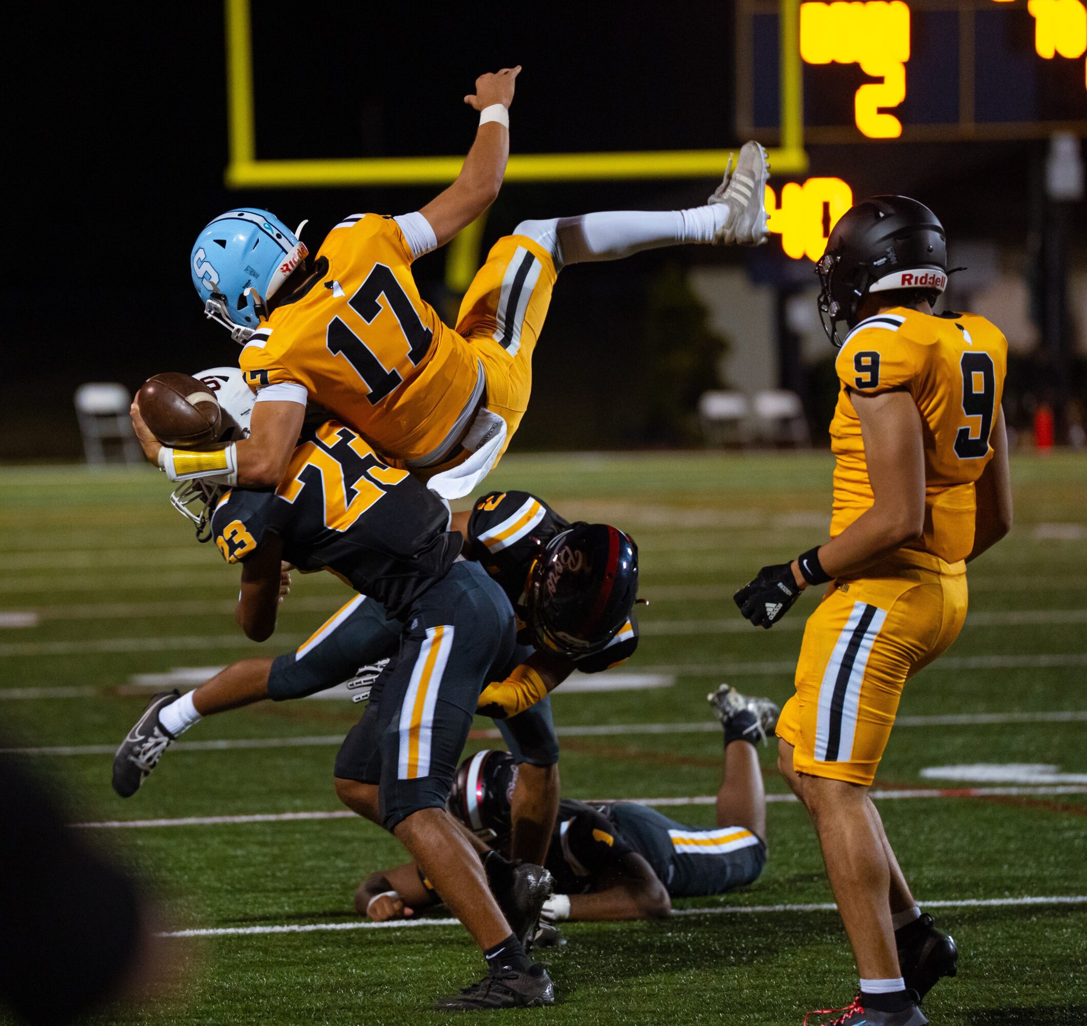 Football players in yellow and black uniforms tackle and block on the field during a game at night.
