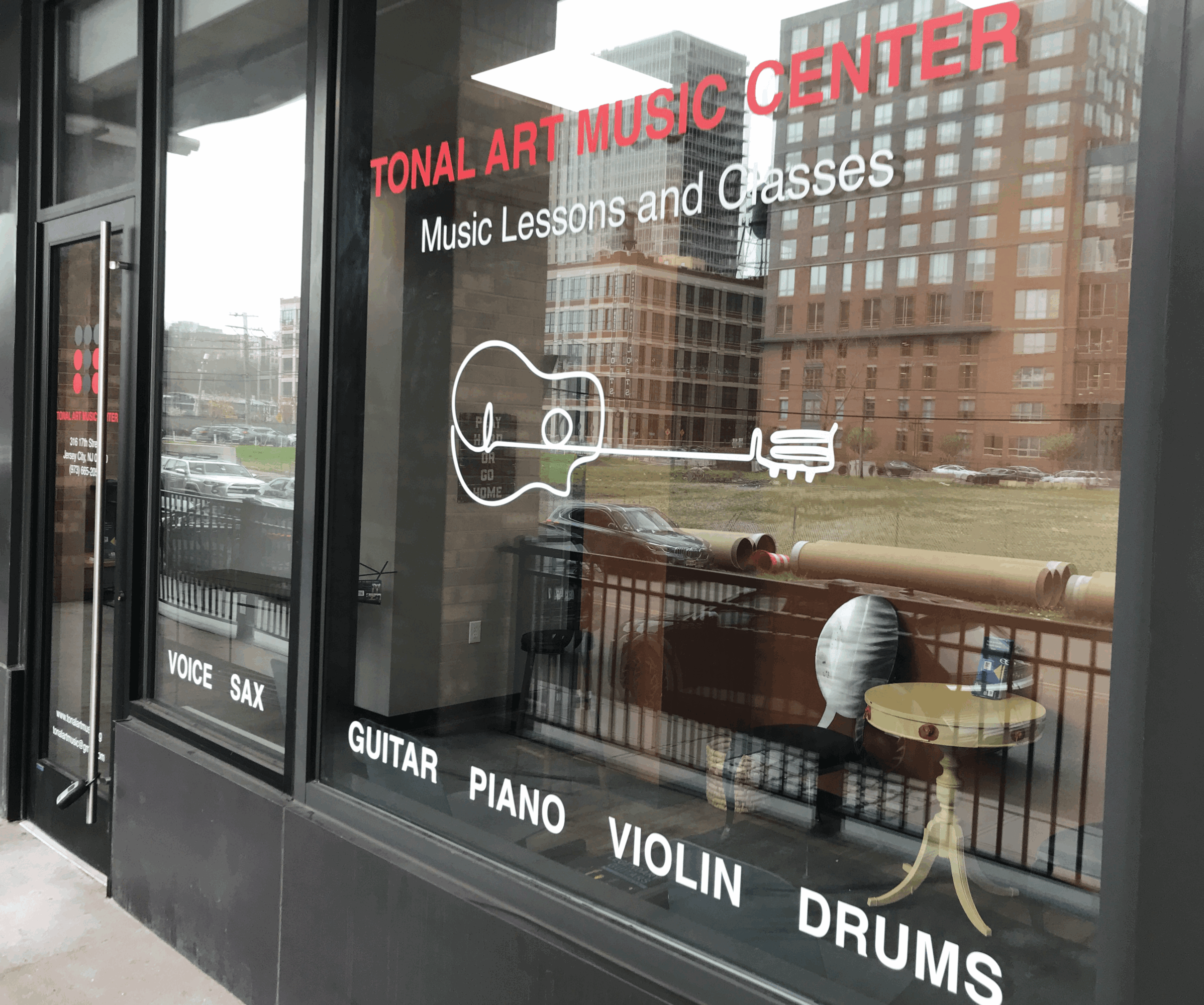 Window display with guitar, piano, violin, and drums illustrations, advertising music lessons at a center, with reflections of buildings and street outside.