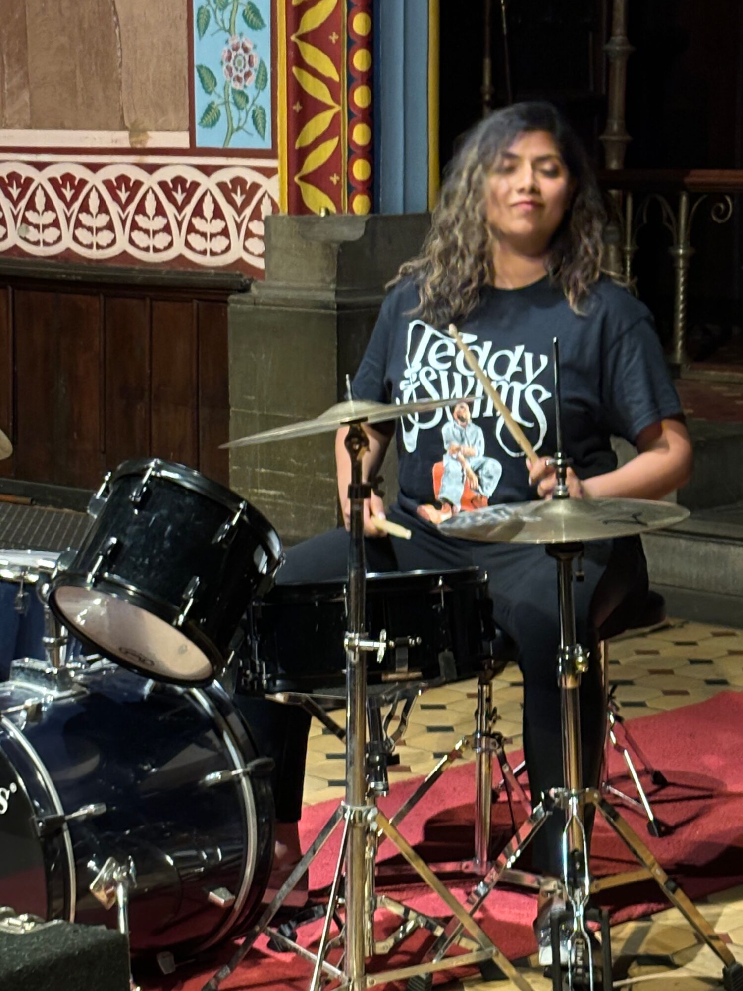 A woman with long, curly hair stands behind a drum set on a stage, holding drumsticks, with a microphone in front.