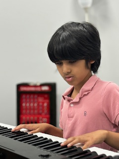 Young boy with black hair playing a keyboard, wearing a pink polo shirt, focused on the keys.