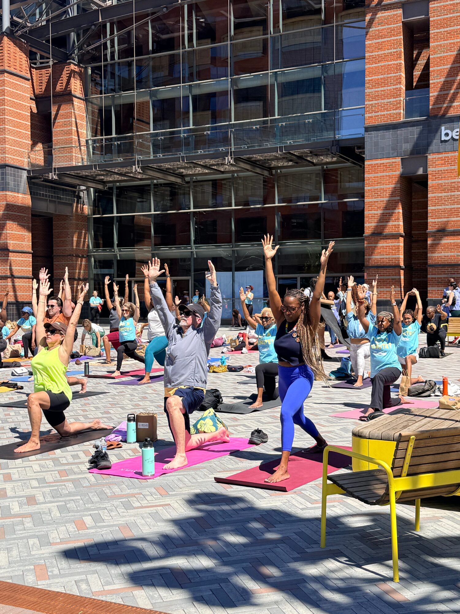 Group of people practicing yoga outdoors on colorful mats in front of a modern building, with a yellow bench nearby.