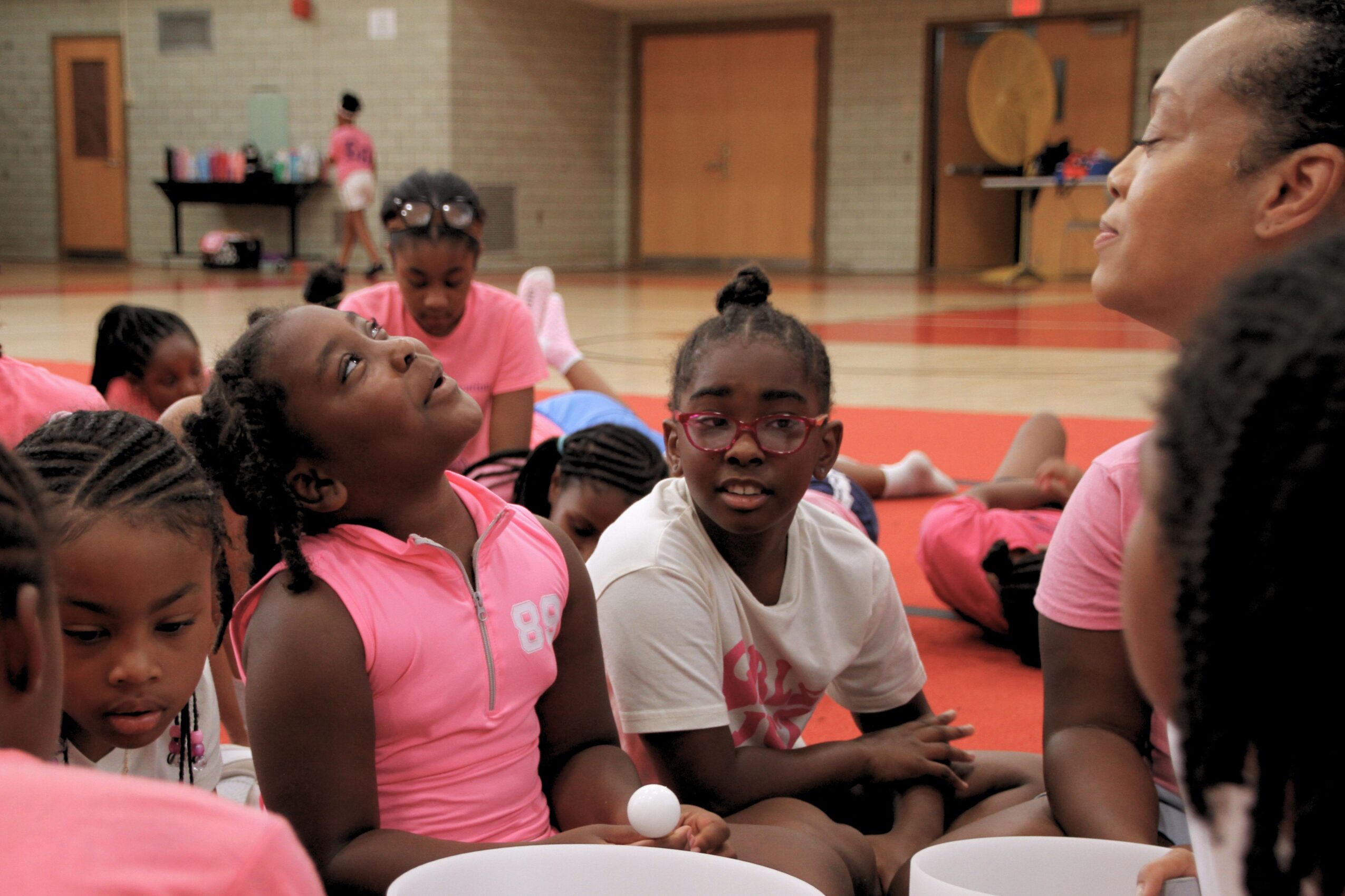 Group of children sitting on gym floor, engaging with an adult, in a gymnasium with wooden floors and equipment.