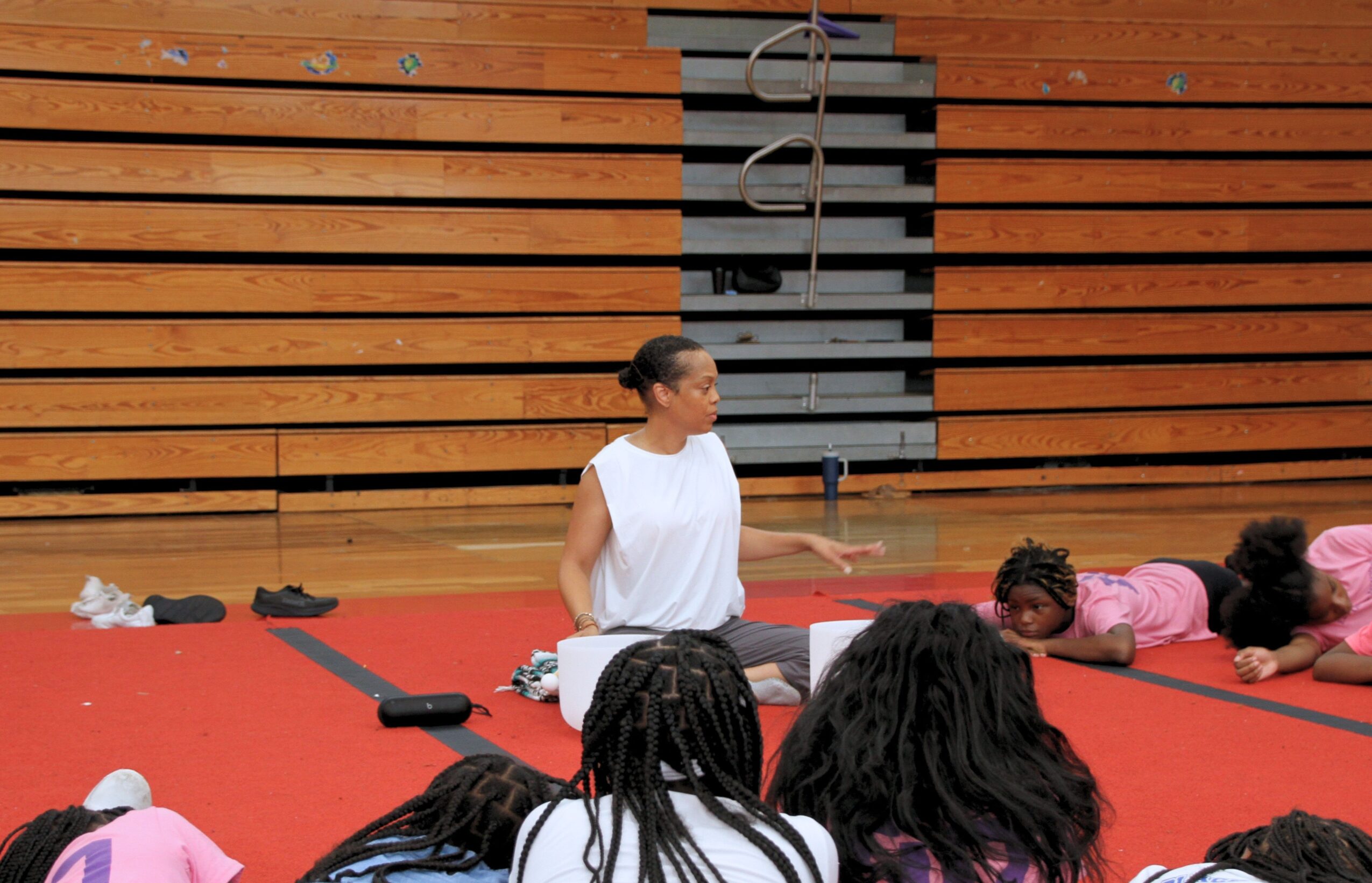 Group of children lying on a red mat, with a woman sitting in the center, in a gymnasium with wooden walls.