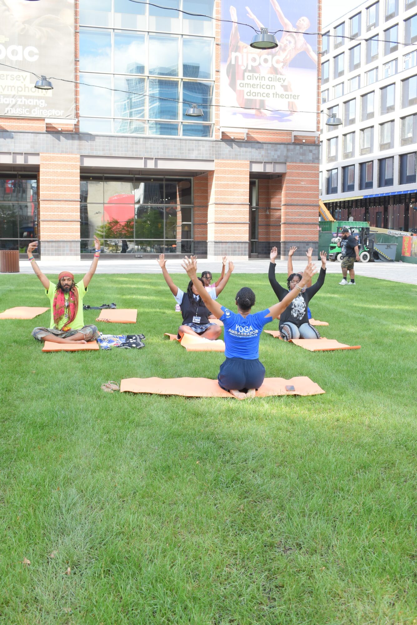 Five people practicing yoga outdoors on a grassy area in an urban setting, sitting on mats with arms raised, facing away from the camera.