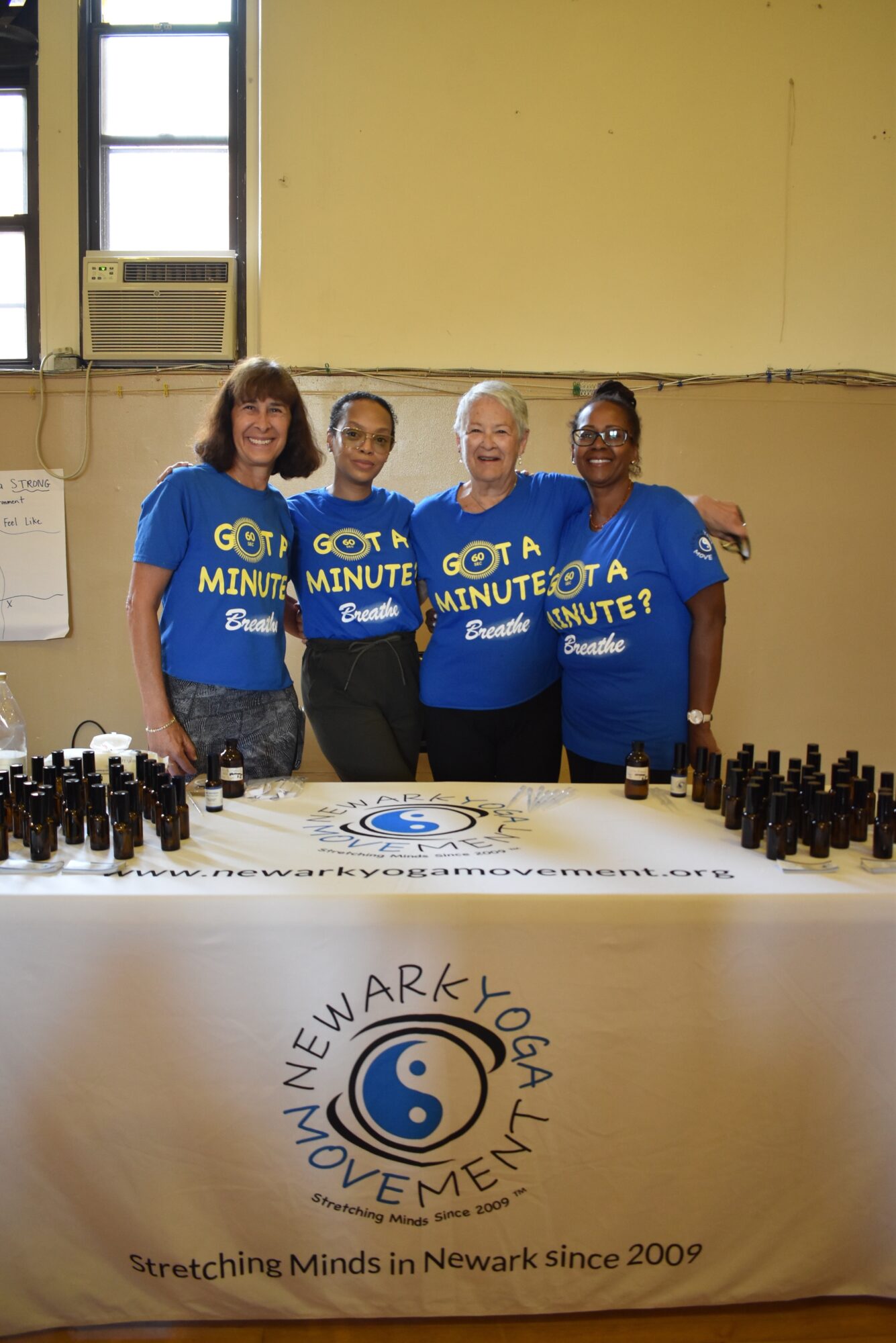 Four women in blue shirts stand behind a table with bottles, smiling indoors. A logo for Newark Yoga Movement is visible on the tablecloth.