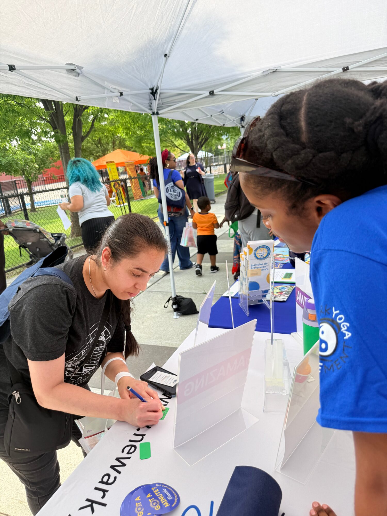 Two women at an outdoor event under a white canopy, one signing a paper, others in background, trees and playground visible.