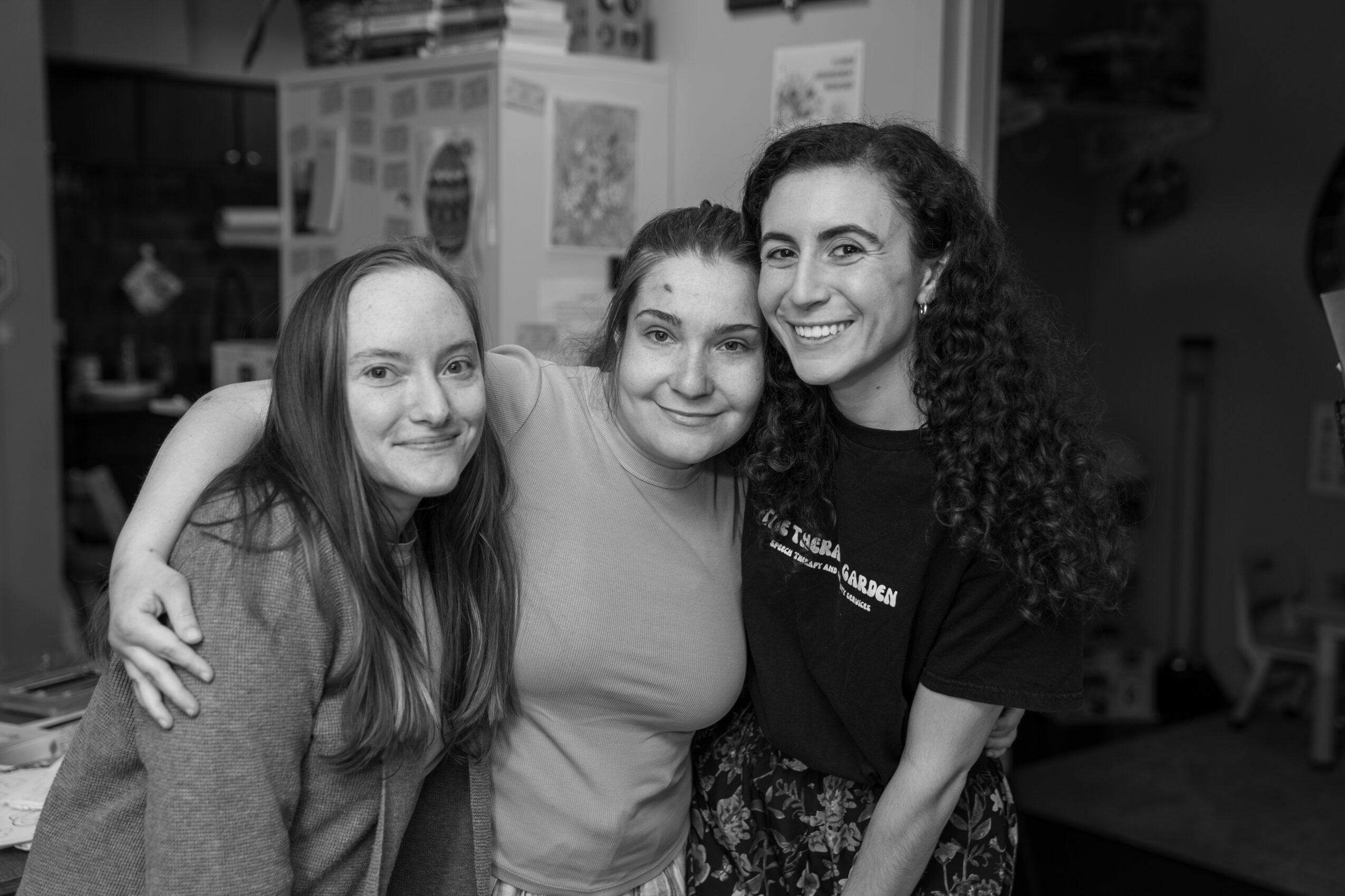 Three women standing close together, smiling, with arms around each other in a room, black and white photo.
