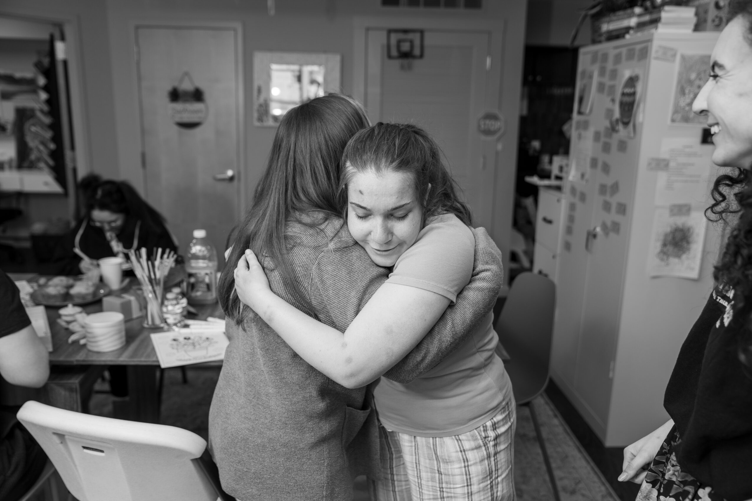 Two women hugging in a kitchen, with a third woman smiling nearby, and a table with various items in the background.