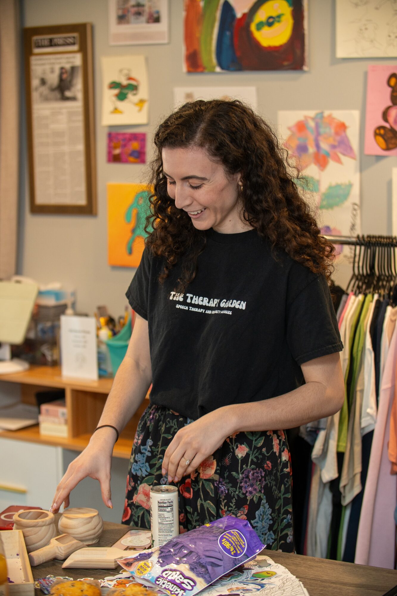 Young woman with curly hair smiling, standing at a table with art supplies, in an art studio with colorful artwork on walls.