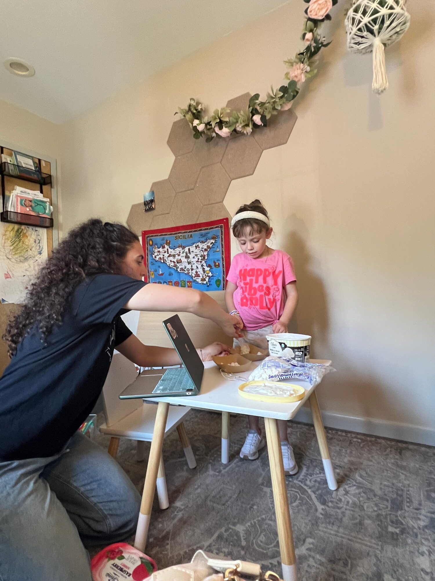 Child in pink shirt and face mask standing near a decorated wall, with a woman sitting at a table with a cake and a laptop.