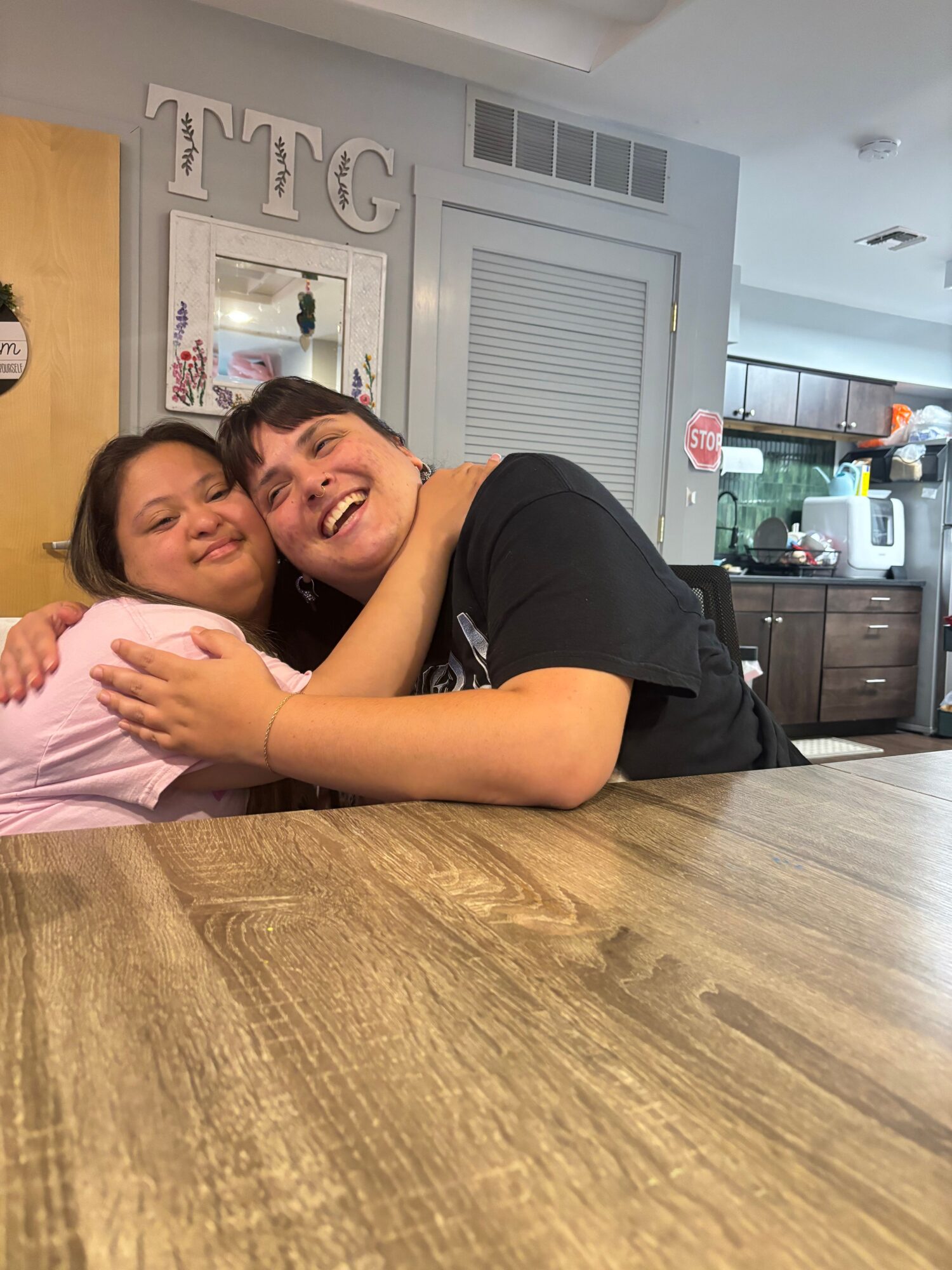 Two women lying on a wooden floor, smiling and hugging each other in a kitchen.