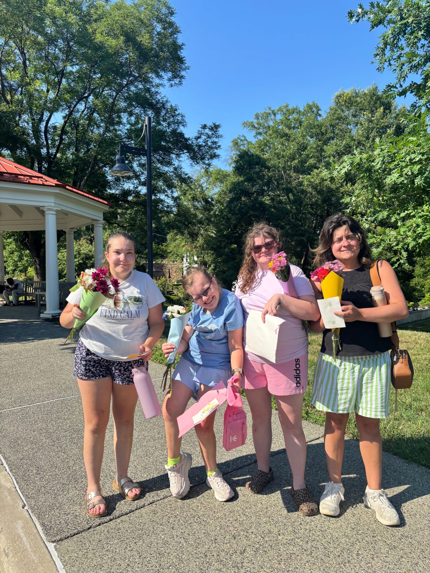 Four young girls standing outdoors on a sunny day, holding flowers and papers, with trees and a bus stop in the background.